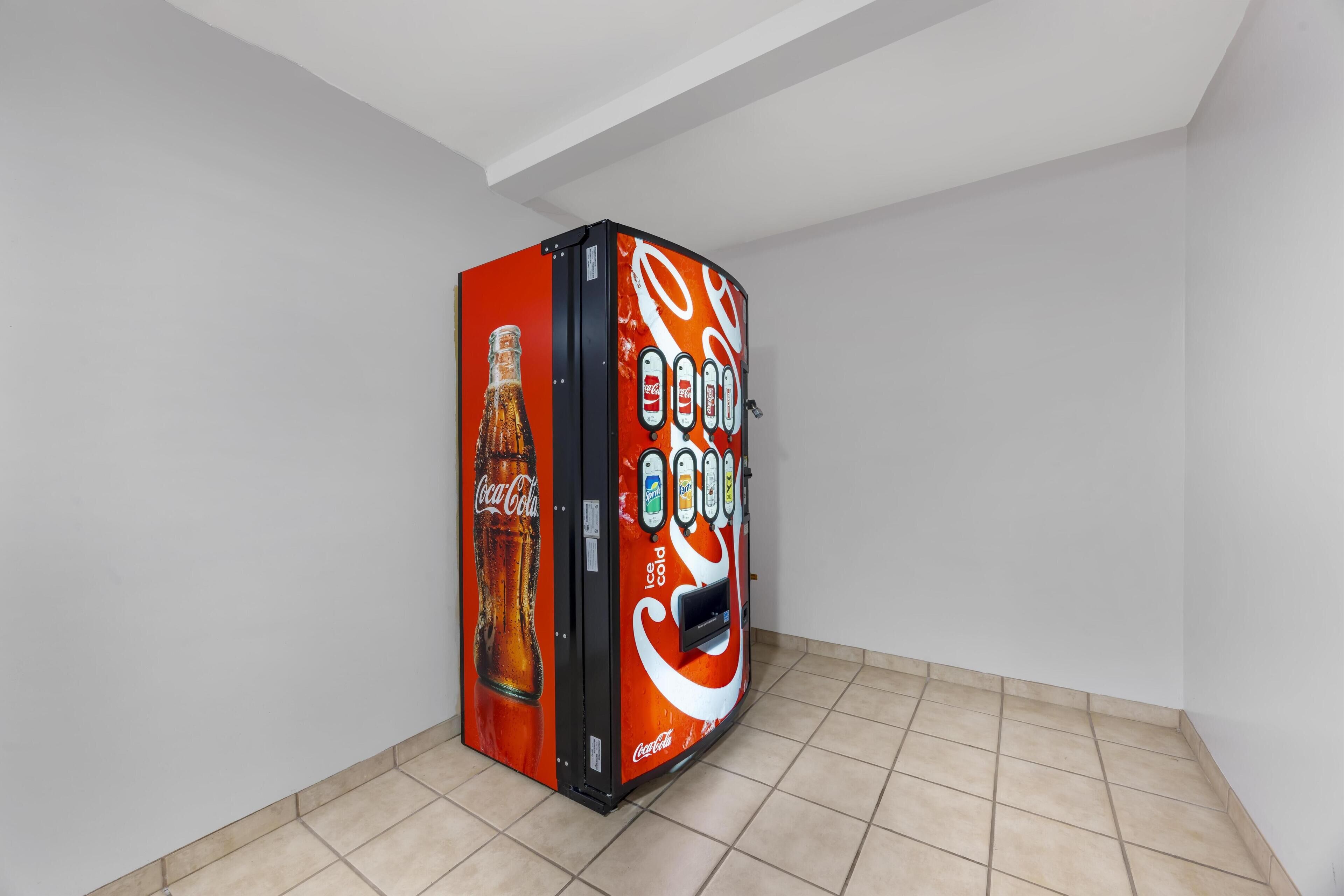 A Coca-Cola vending machine in a corner of a room with tiled flooring and white walls.