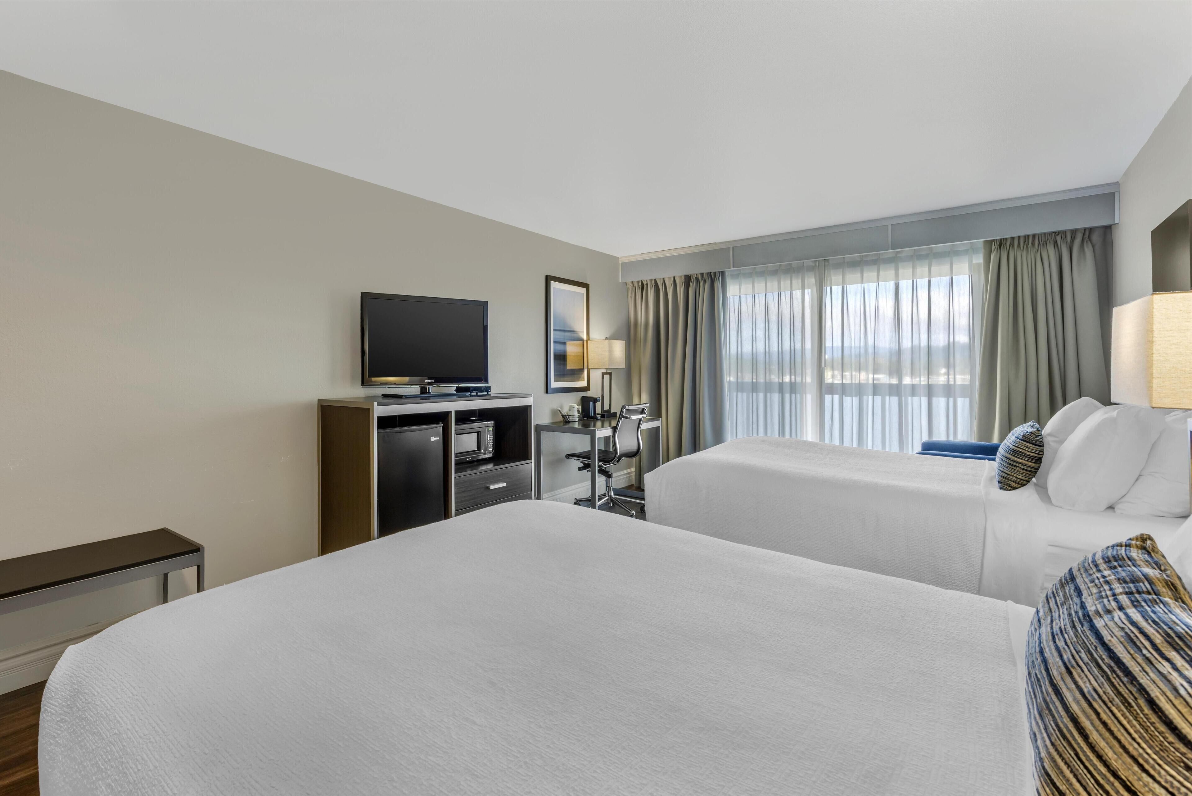 Habitación de hotel con dos camas, un escritorio, un televisor de pantalla plana, un mini refrigerador y una ventana con cortinas.