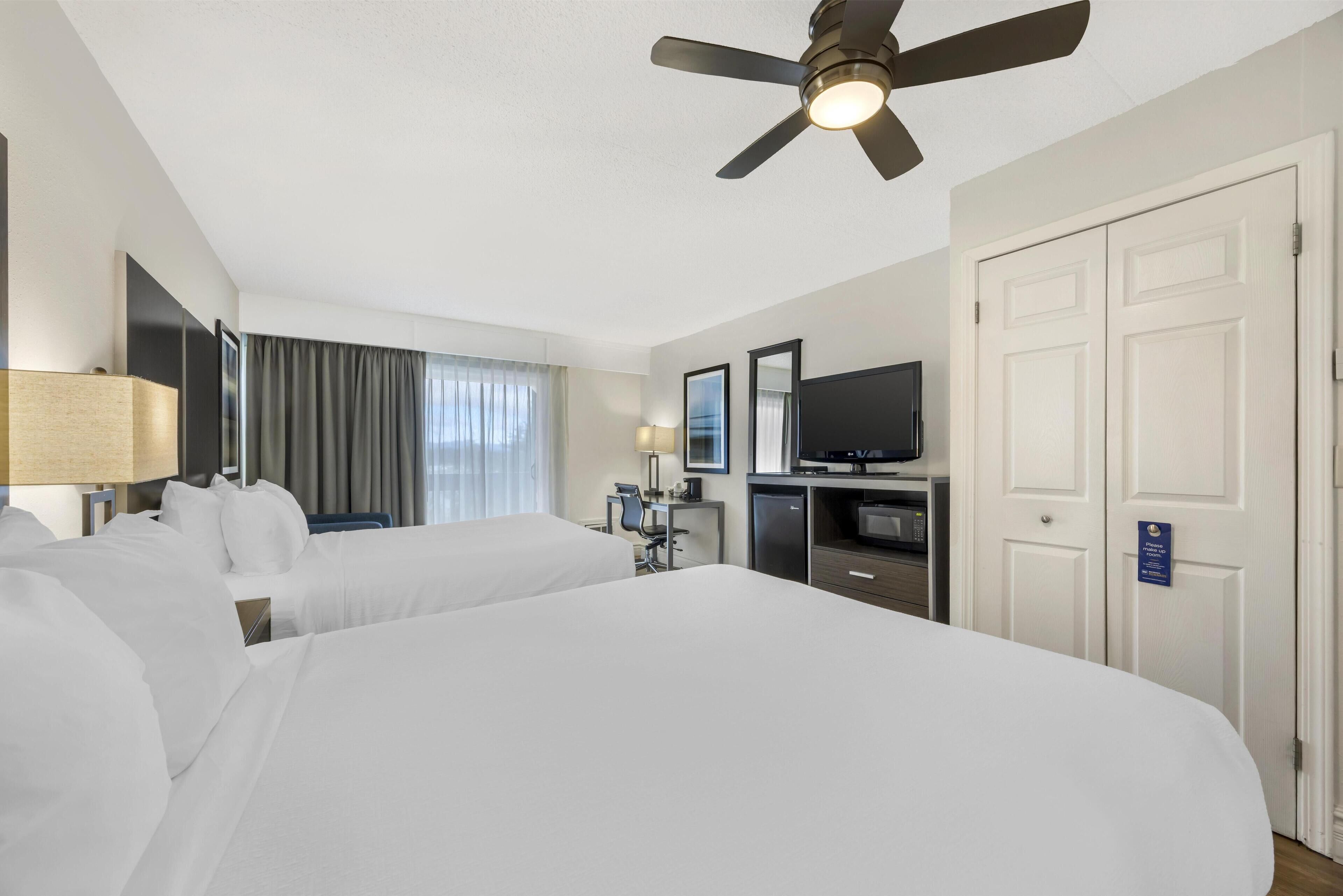 A hotel room with two beds, a desk, a television, a ceiling fan, and a door with a sign.