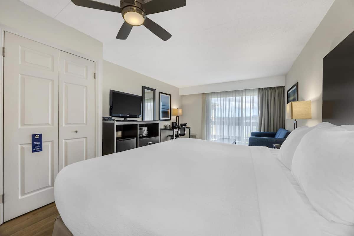 Habitación de hotel con una cama grande, un escritorio con computadora, televisor y una zona de estar con una silla y sofá.