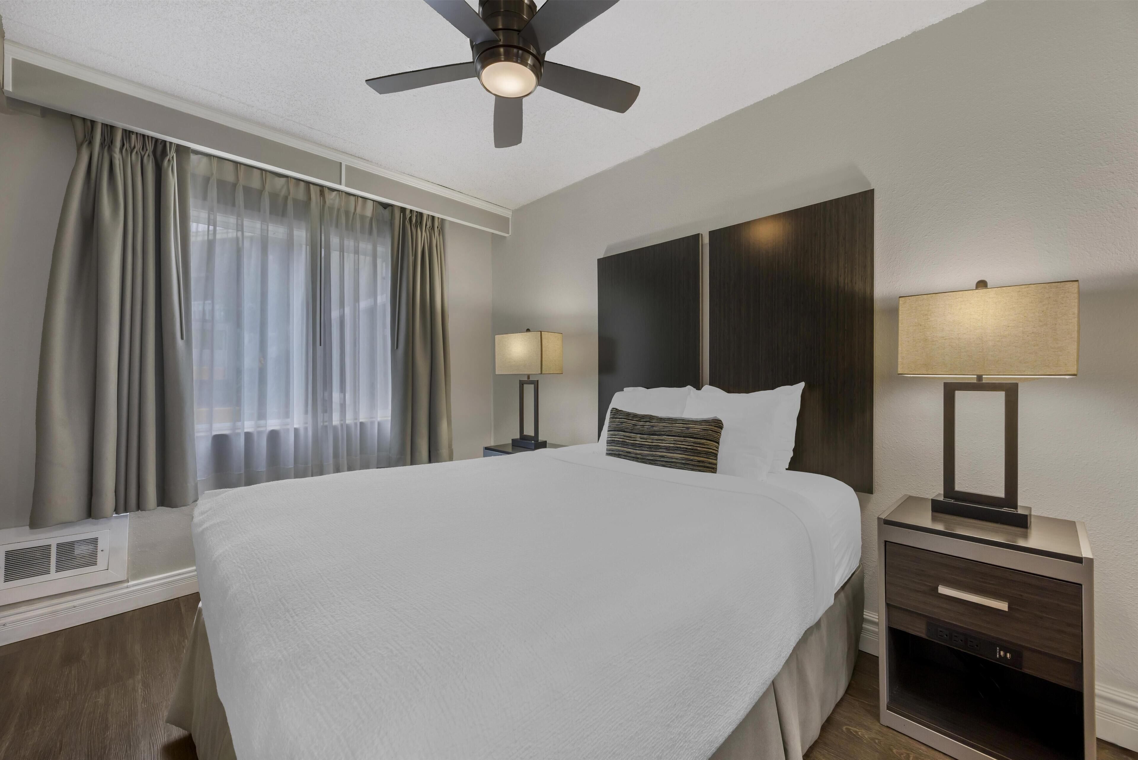 Una habitación de hotel con una cama grande, dos mesitas de noche con lámparas, un ventilador de techo y una ventana con cortinas.