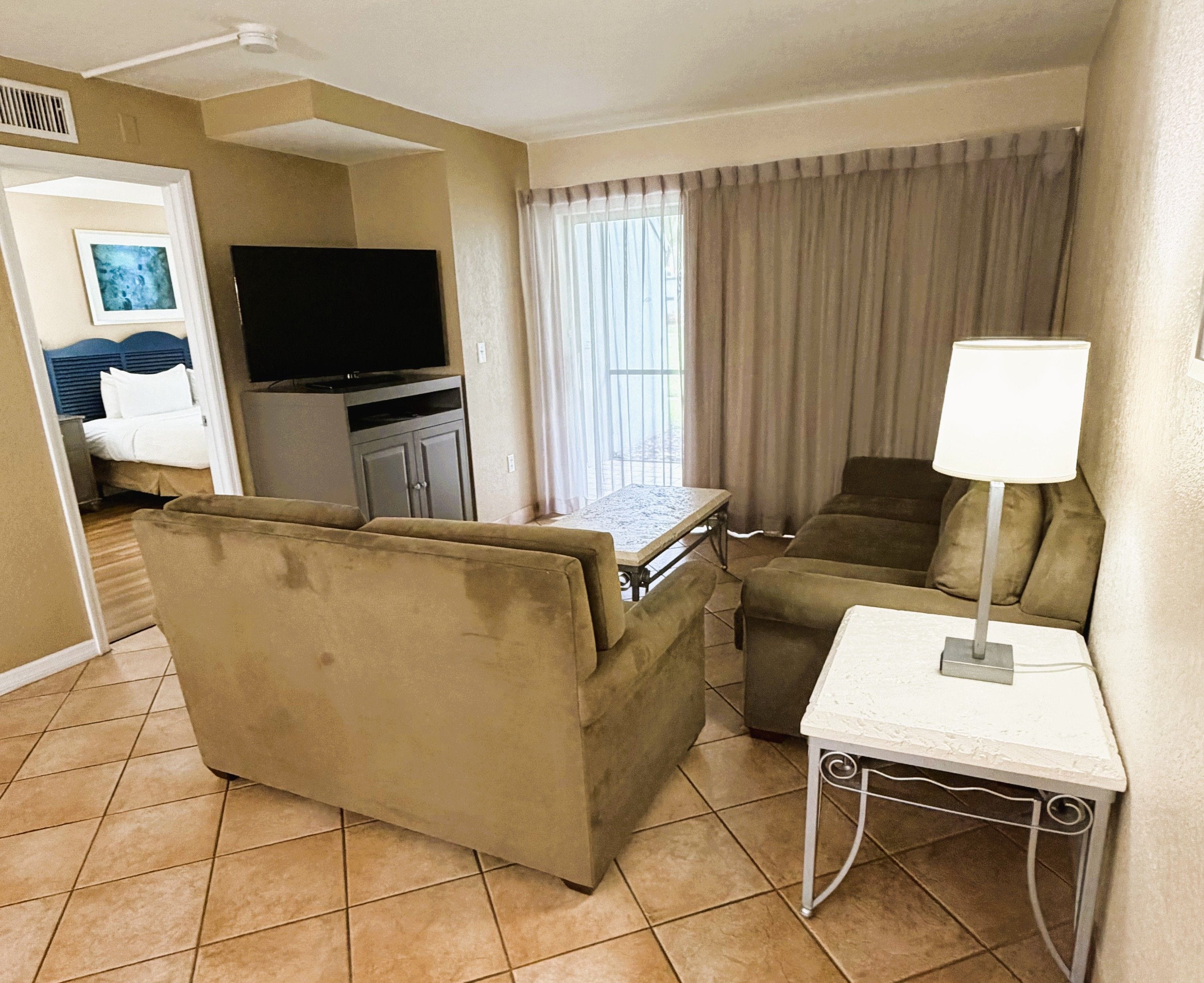 A hotel room with a sofa, a small table with a lamp, a television, and a window with curtains.