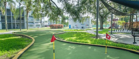 A miniature golf course with a green putting surface, two red flags marking the holes, and a playground in the background.