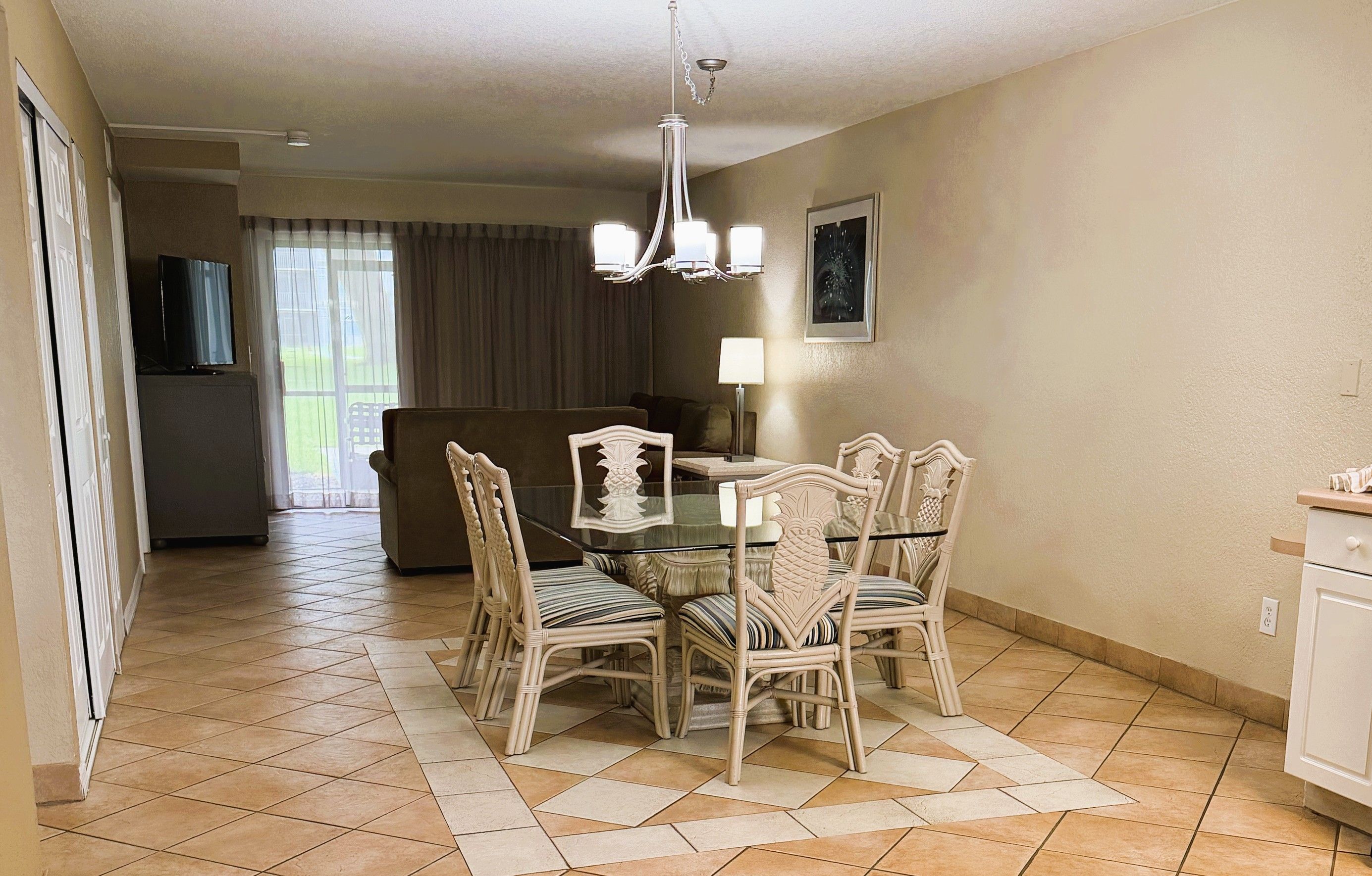 A dining area with a glass table surrounded by wicker chairs, a chandelier, and a view of a living room with a sofa and curtains.
