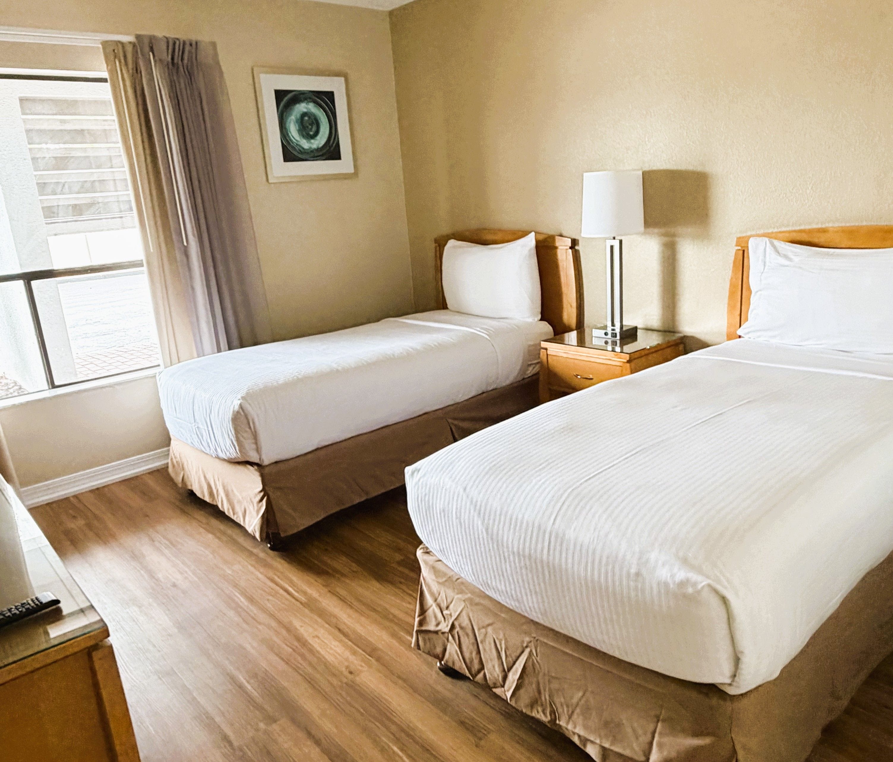 A hotel room with two beds, a window with curtains, a bedside table with a lamp, and a wall-mounted picture.