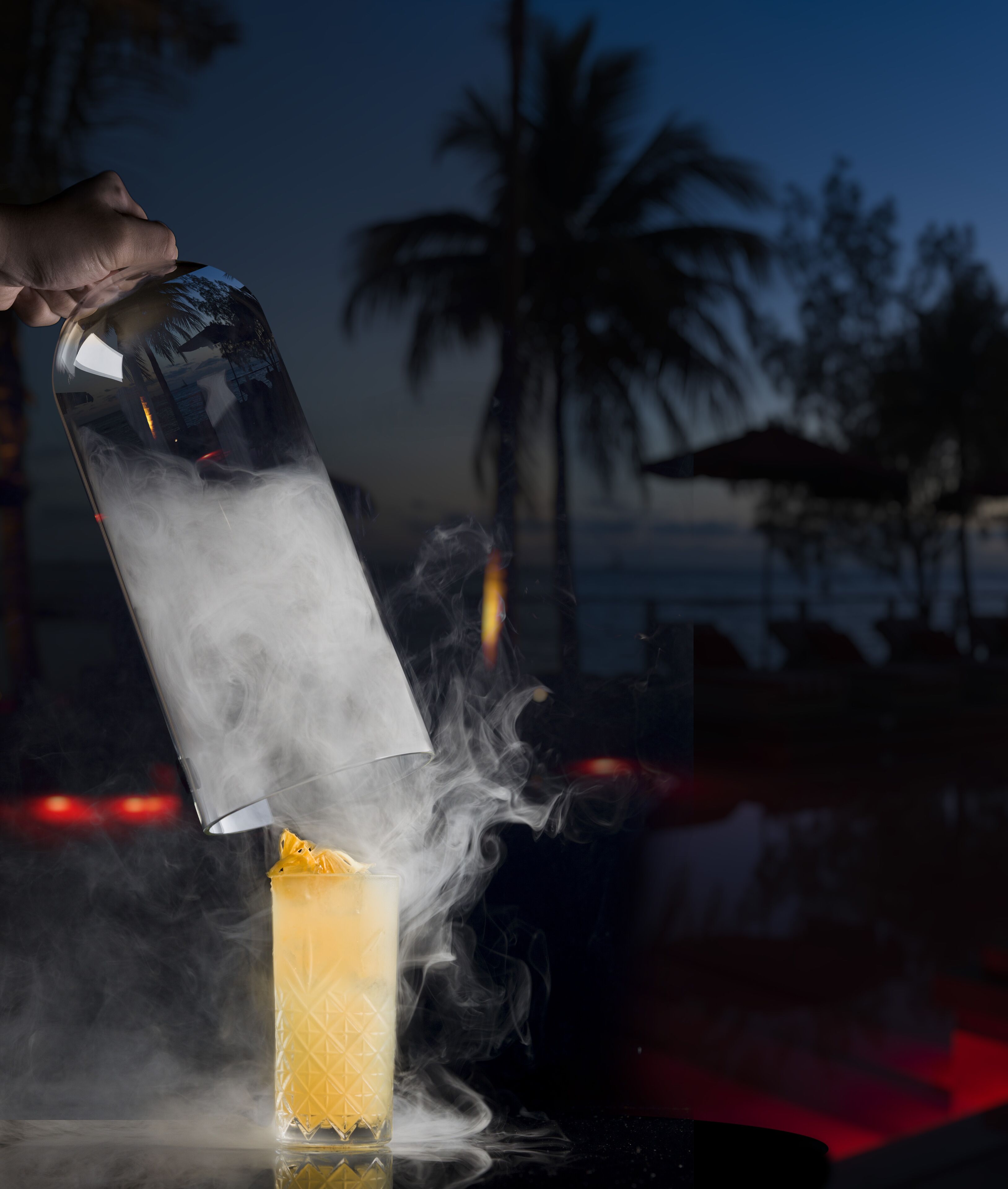 A cocktail being poured into a glass with smoke effects, set against a beach background.