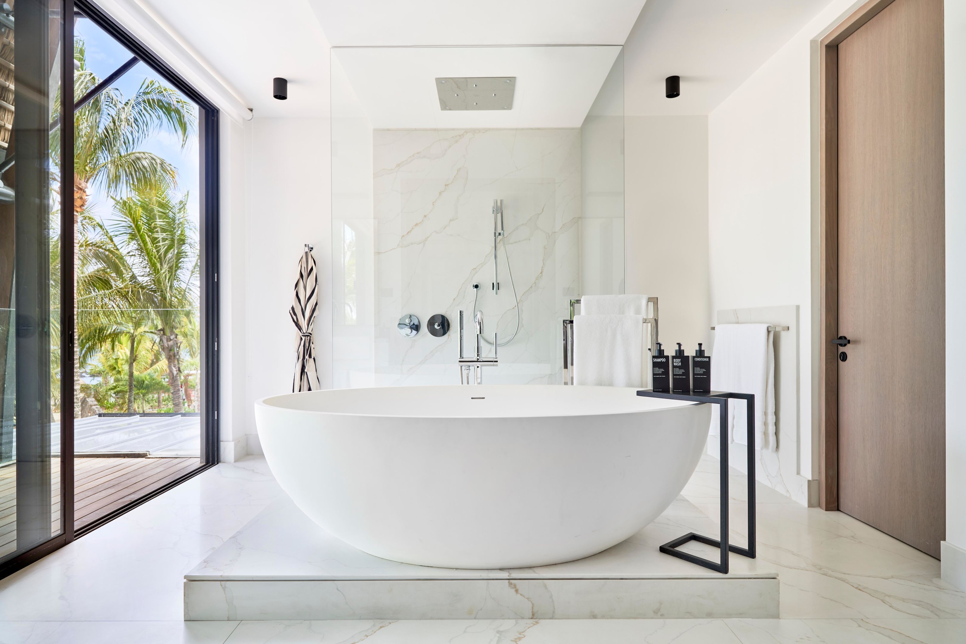 A modern bathroom with a freestanding bathtub, a glass shower enclosure, and a view of palm trees.
