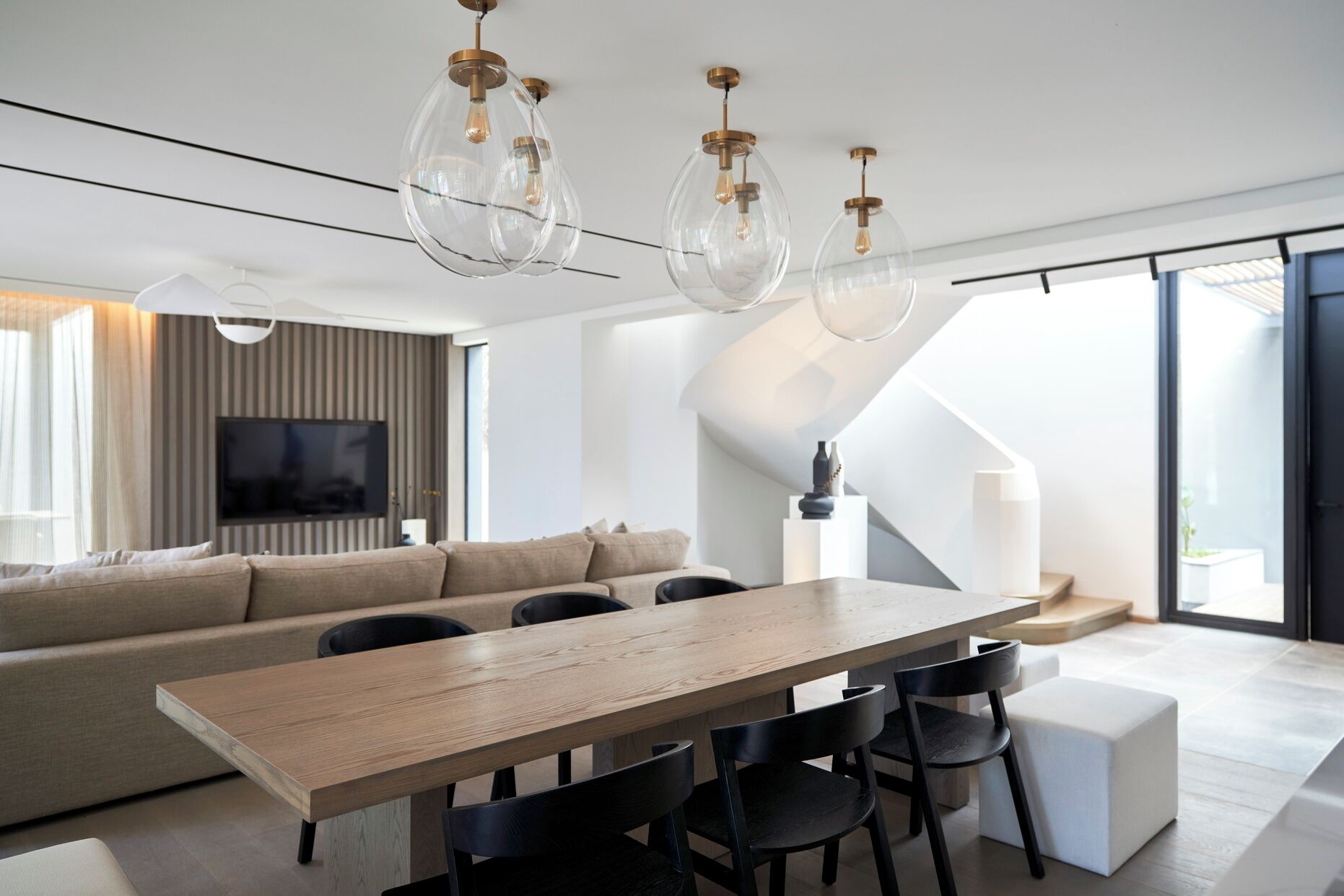 Una sala de estar moderna con una mesa de comedor de madera, sillas negras, un sofá beige y un televisor montado en la pared.