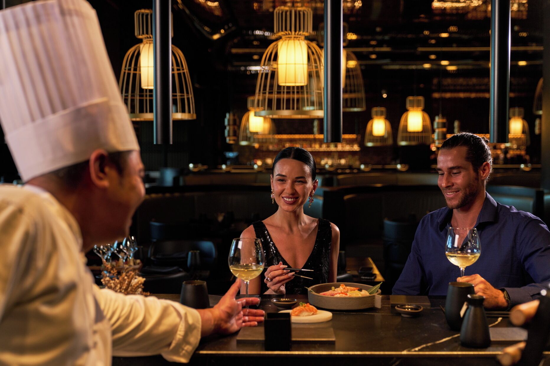A restaurant interior with tables, hanging light fixtures, and a chef in a white hat.