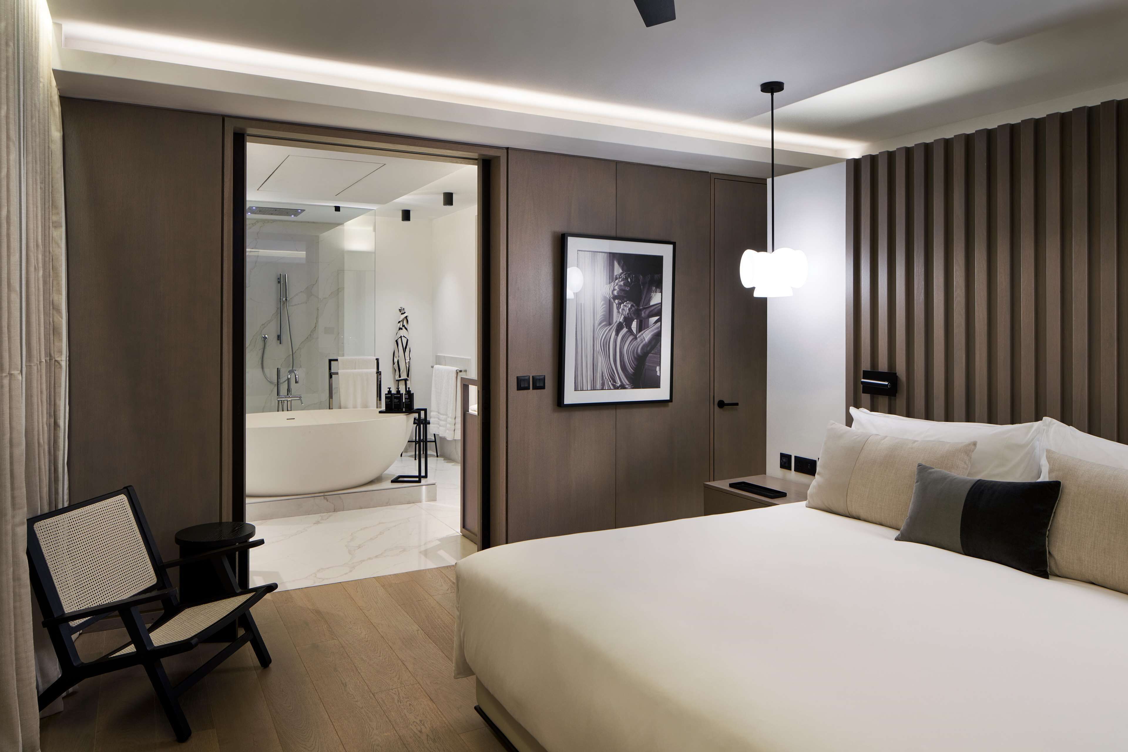 Ein modernes Hotelzimmer mit einem großen Bett, einem kleinen Nachttisch, einem Sessel, einem gerahmten Kunstwerk, einer Wandleuchte und Blick ins Badezimmer.