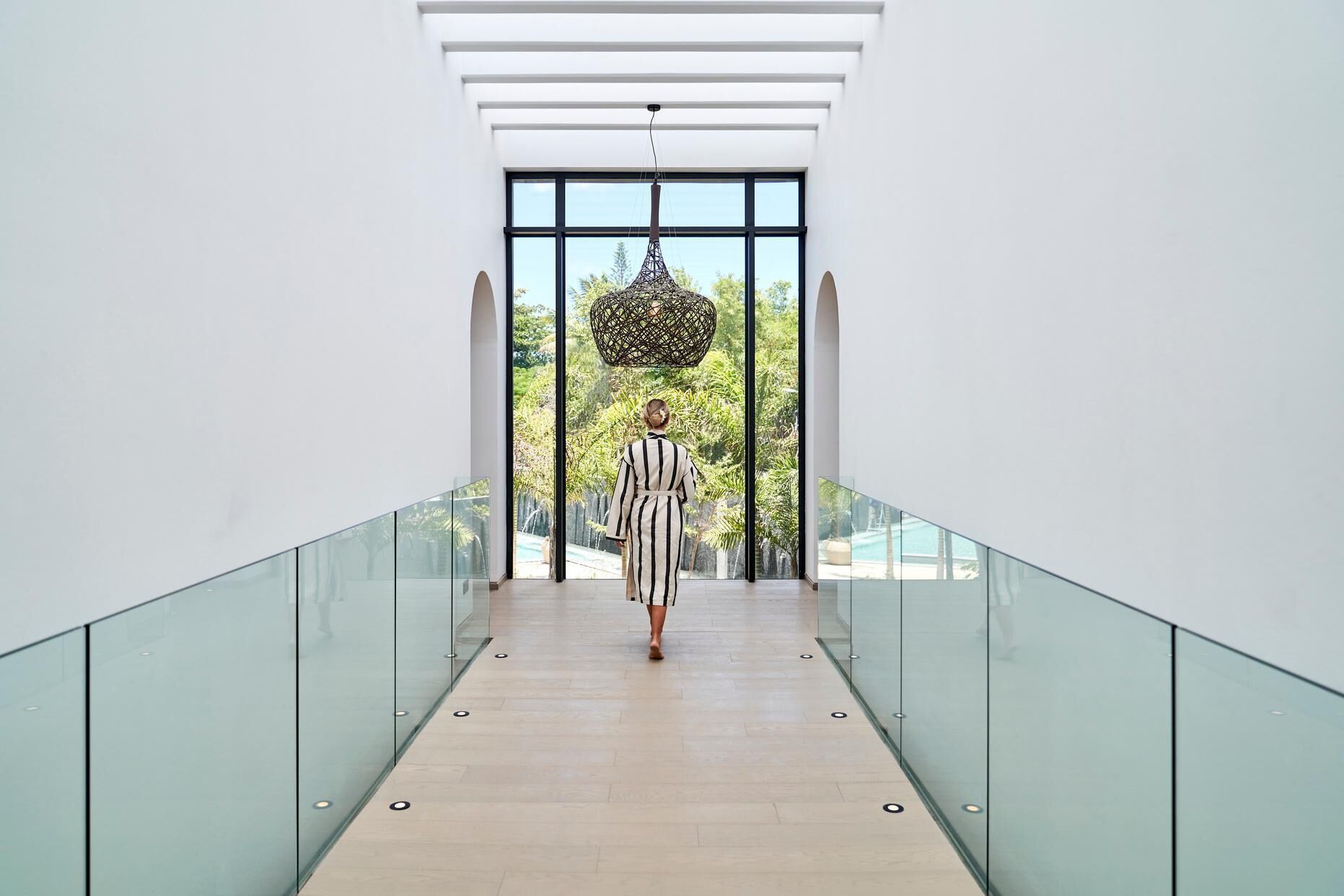 Un interior moderno con barandilla de vidrio, una persona caminando por un pasillo y vistas de la vegetación al exterior.
