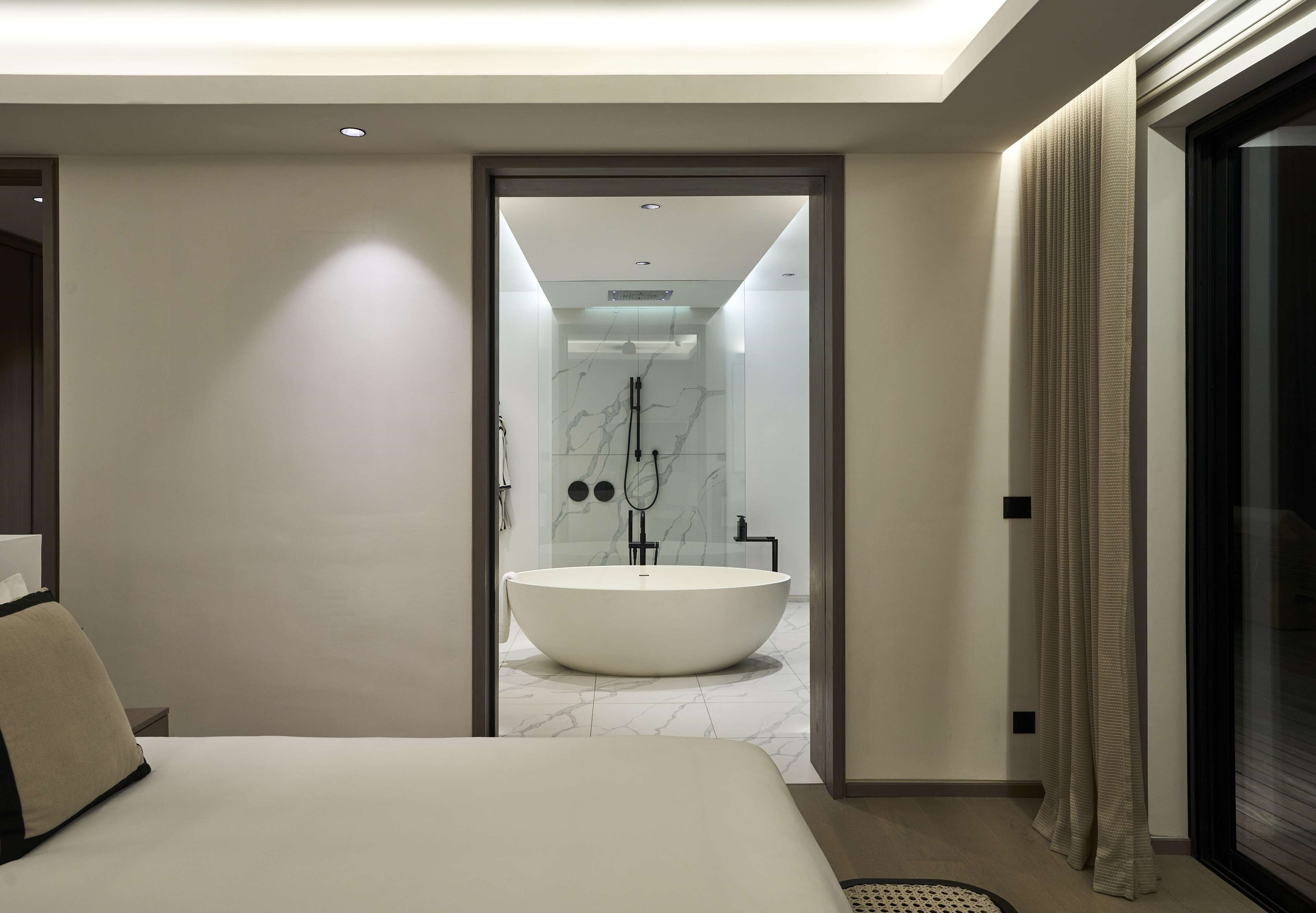 A modern hotel room with a large bed, a bathroom with a freestanding bathtub, and a glass door leading to a balcony.