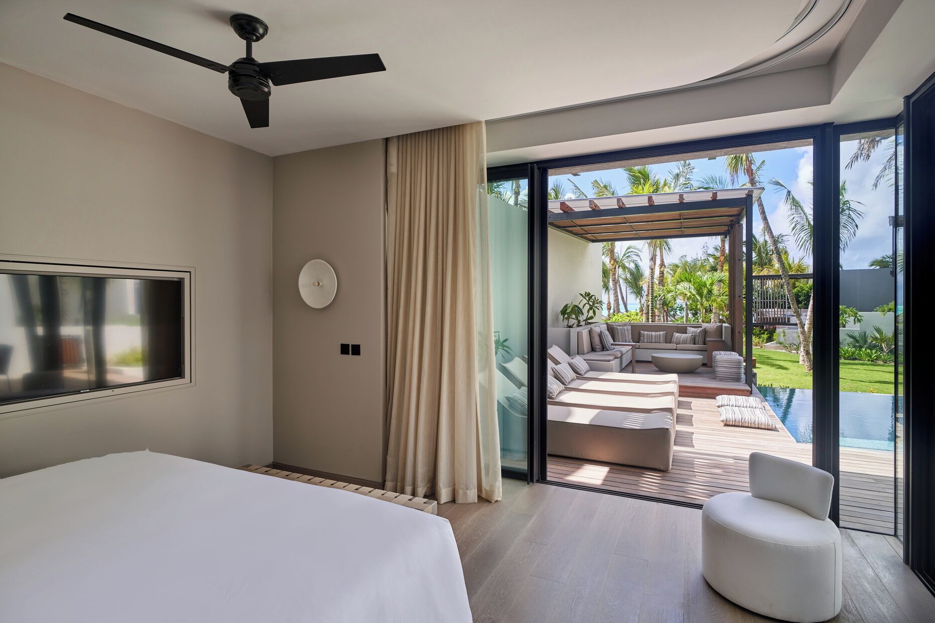 Una habitación de hotel moderna con una cama, un ventilador de techo, un televisor y un vidrio corredizo que da a una zona de estar exterior con piscina.