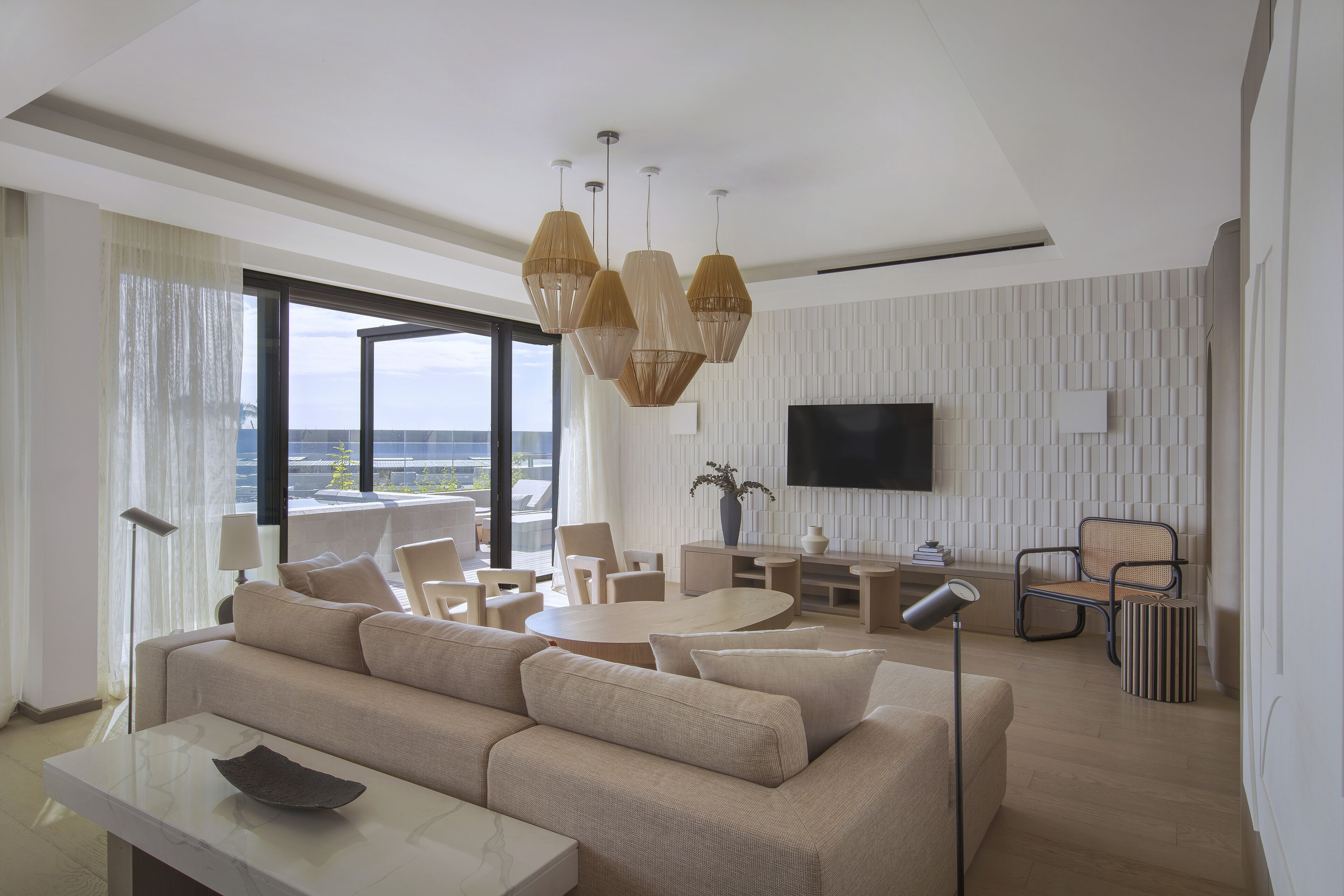A modern living room with a sofa, coffee table, and dining area. There is a TV mounted on the wall and a large window providing a view of the outdoors.