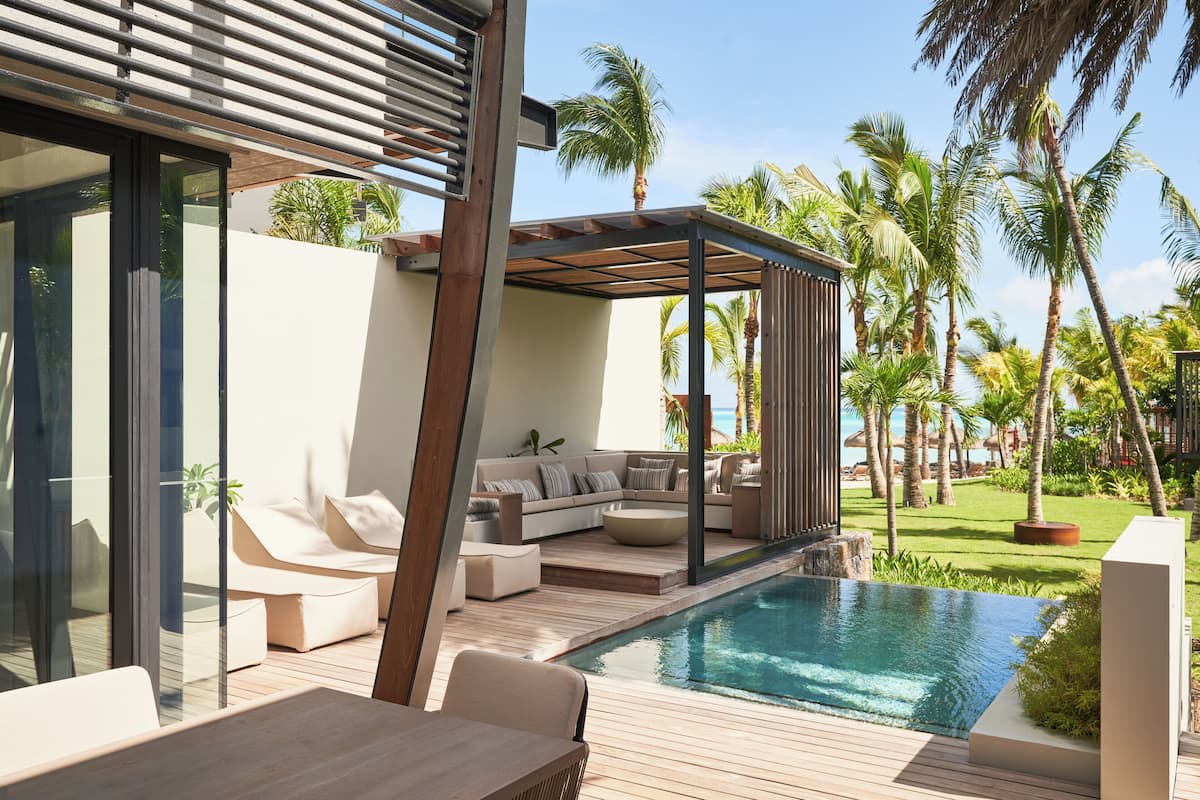 Een zwembad buiten met een modern design, een houten terras, een overdekte zithoek en weelderige beplanting, waaronder palmbomen.