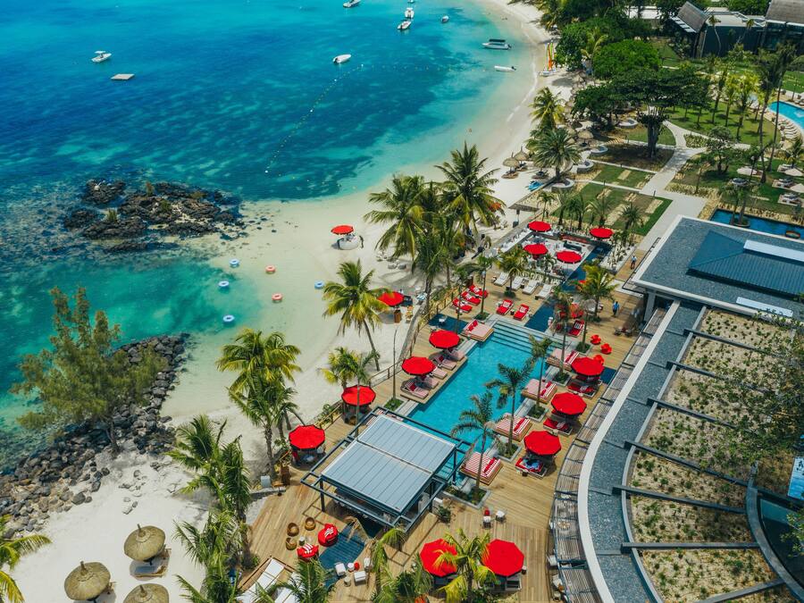 An aerial view of a resort with a beach, a pool area with umbrellas, and a clear view of the ocean.