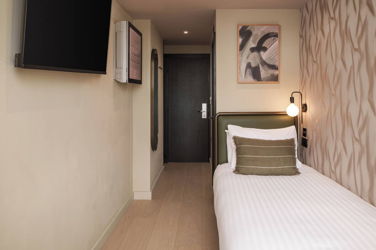 A hotel room with a bed, a wall-mounted TV, a framed picture, a door, and a light fixture.