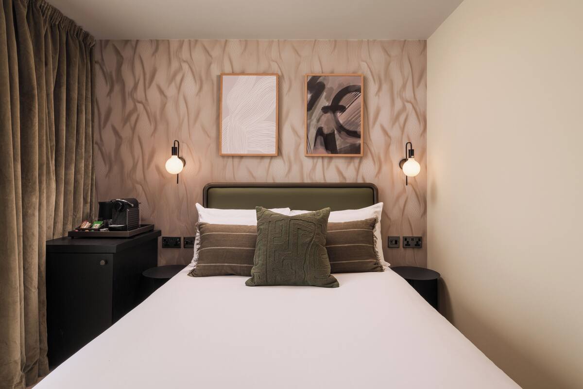 A neatly arranged hotel room with a bed, two bedside lamps, a nightstand with a coffee maker, and a wall with textured wallpaper and two framed artworks.