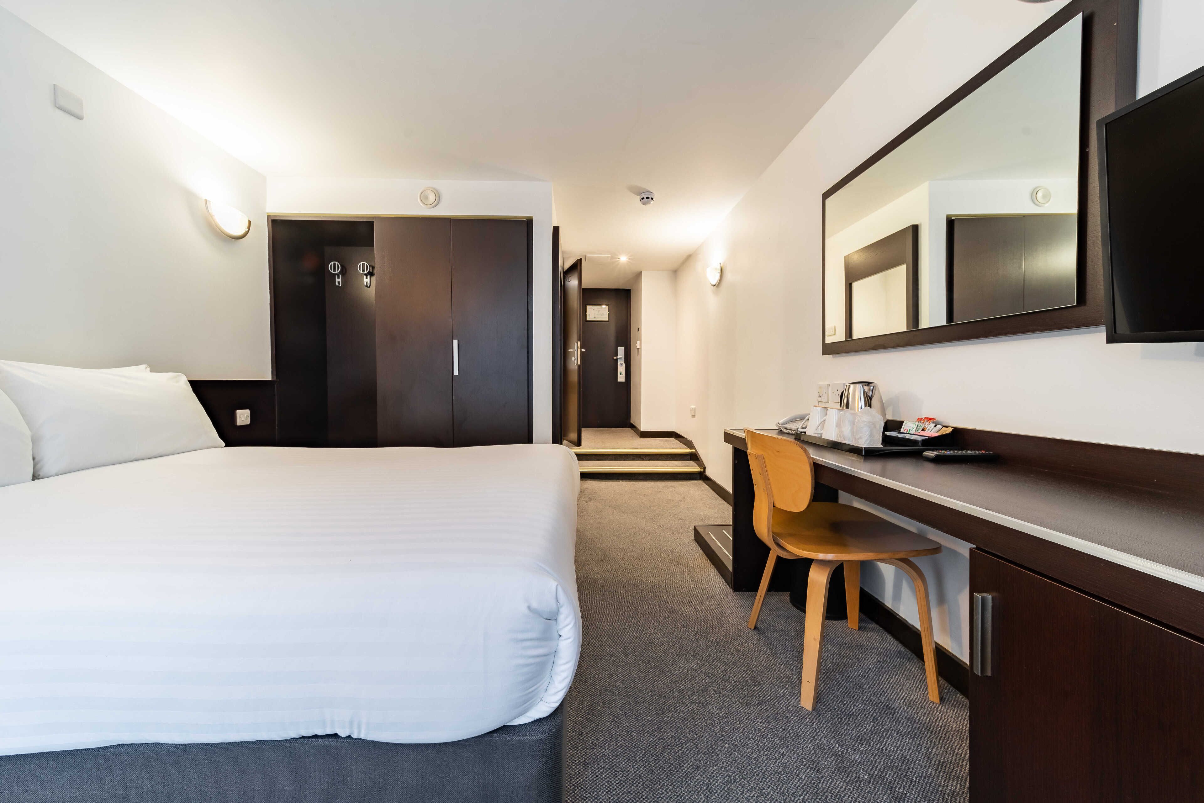 A hotel room with a bed, a desk with a chair, a TV, a mirror, and a door leading to a hallway.
