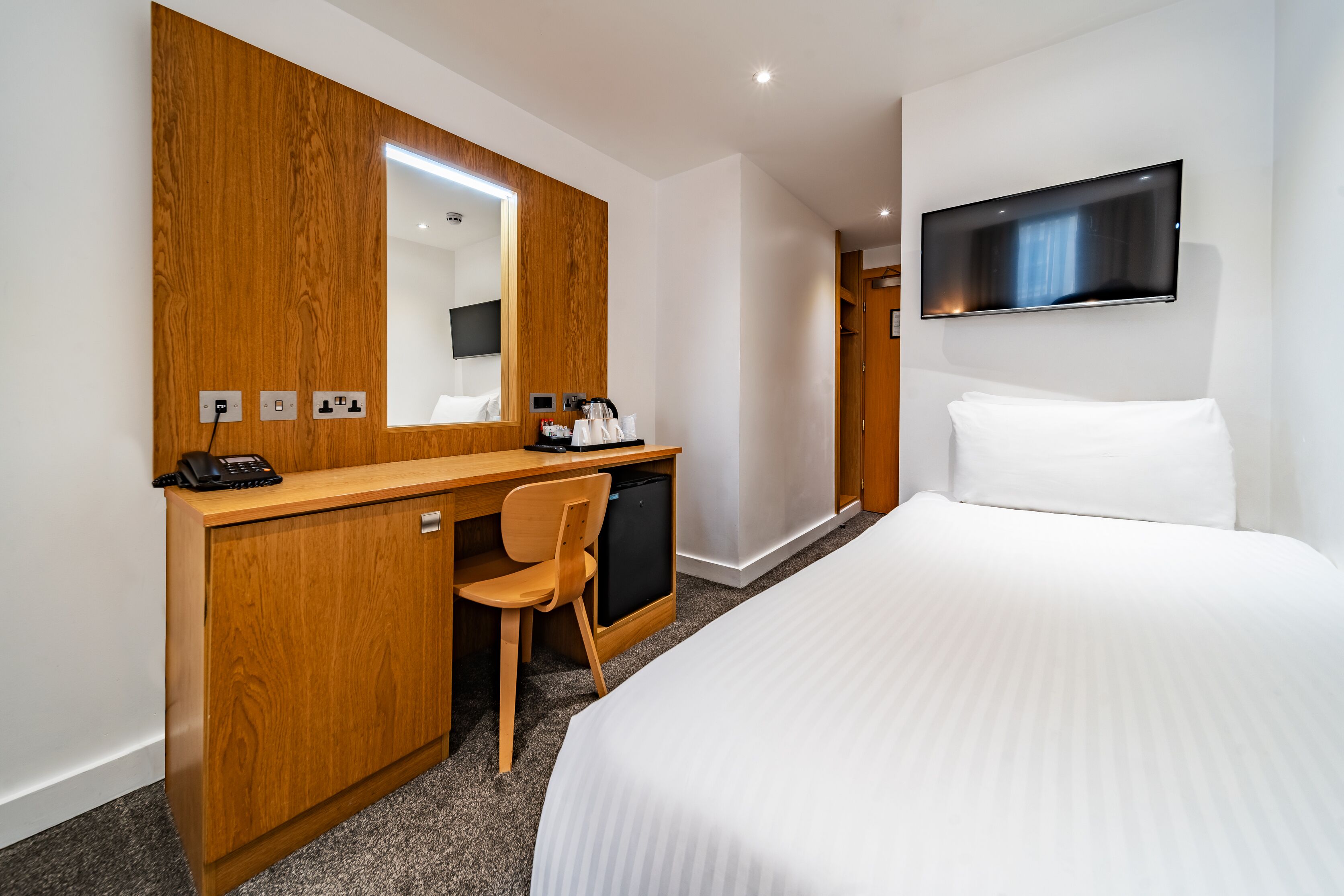 Une chambre d’hôtel avec un bureau en bois, une chaise, un lit avec du linge de maison blanc, un téléviseur fixé au mur et un miroir au-dessus du bureau.