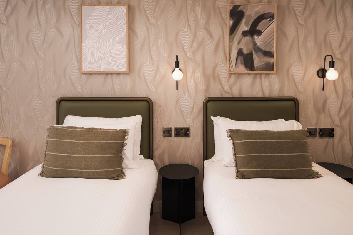 A hotel room with two beds, each with a headboard, a round black side table, and wall-mounted light fixtures.