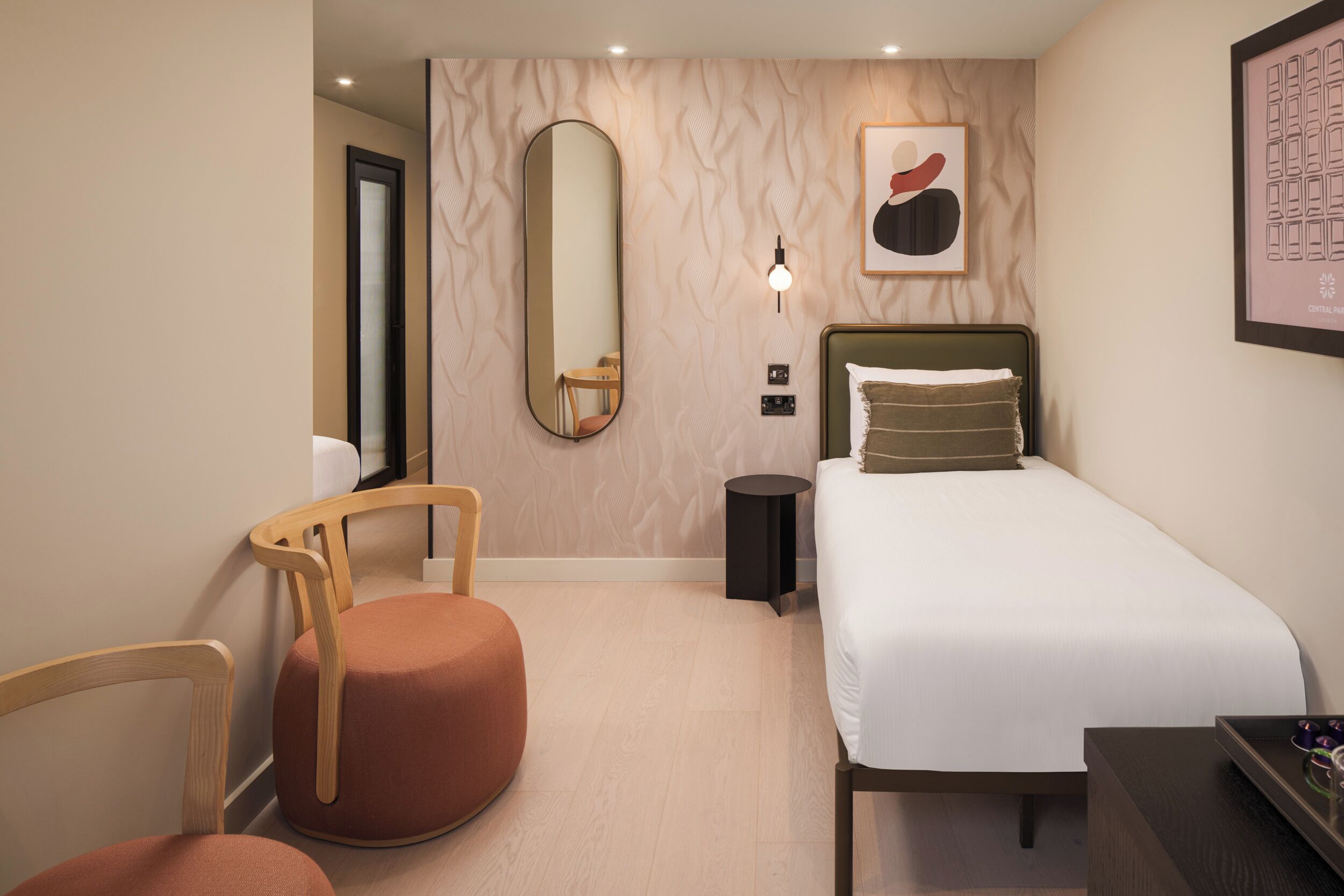Habitación de hotel con una cama, una silla redonda de madera, una mesita redonda pequeña, un espejo adosado a la pared y una obra de arte enmarcada.