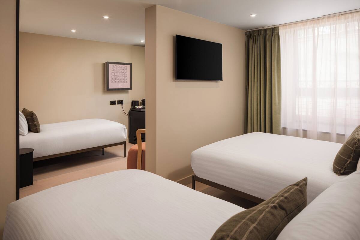 A hotel room with two beds, a flat-screen TV, a framed picture on the wall, and a window with sheer curtains.