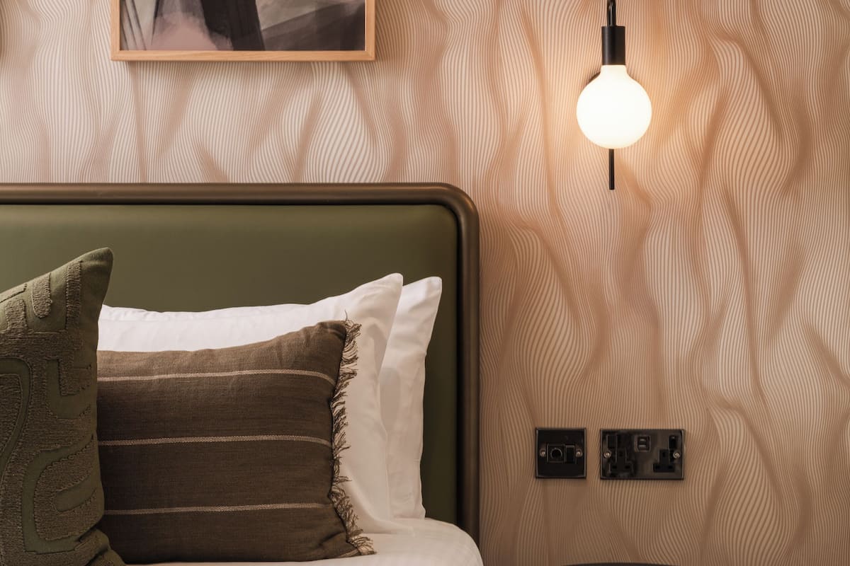 A neatly made bed with pillows, a wall-mounted light, and a small black side table.