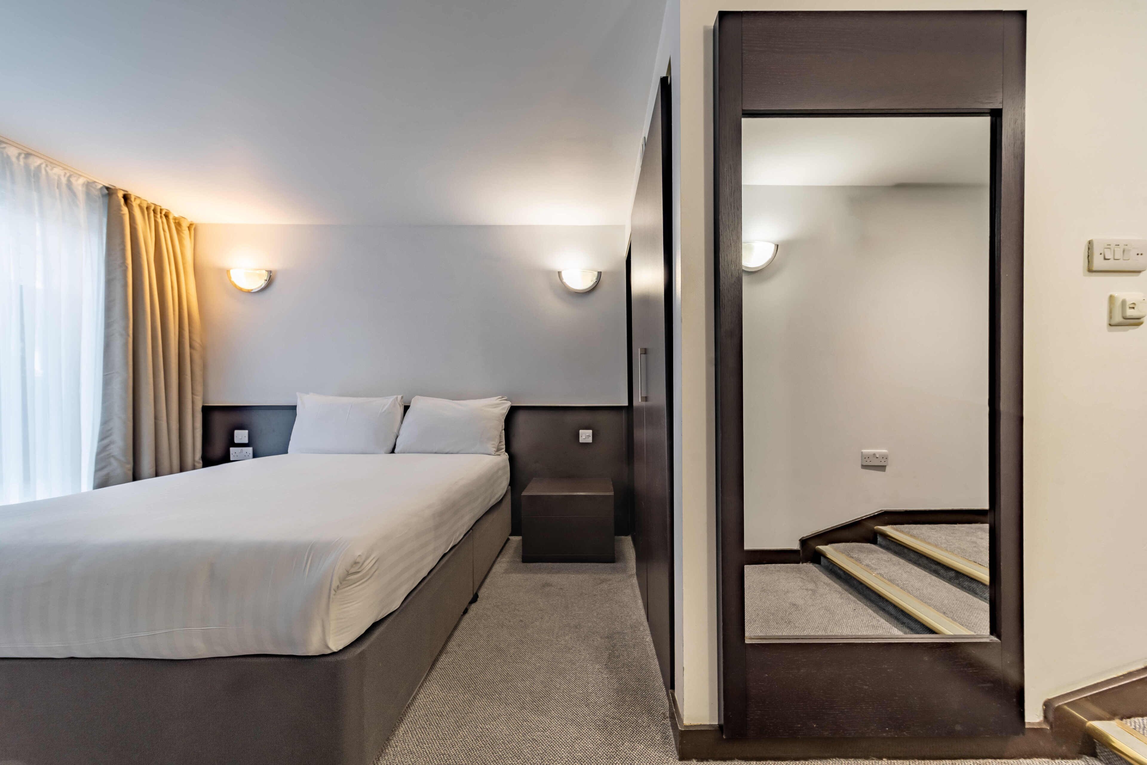 Ein Hotelzimmer mit Bett, Nachttisch und einem Spiegel, der eine Treppe reflektiert.