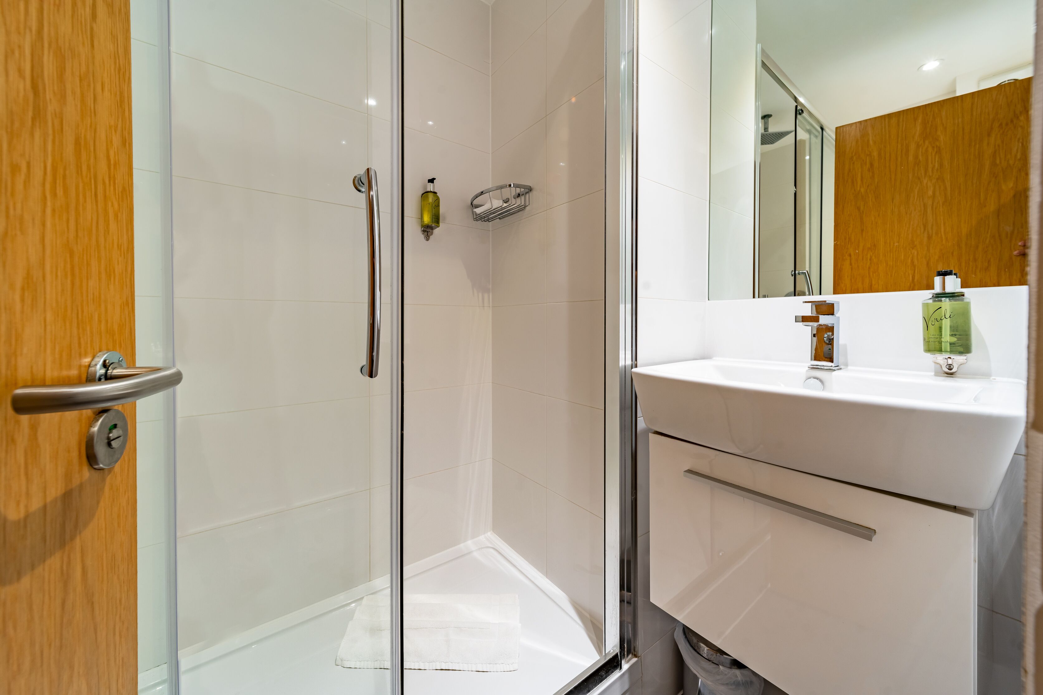 Een moderne badkamer met een douchecabine van glas, een witte wastafel en een houten opbergmeubel.