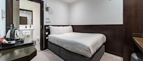 A modern hotel room with a bed, a desk with a kettle, a trash bin, and a mirror.