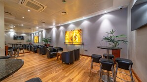 A spacious room with a wooden floor, multiple seating areas including round tables and bar stools, a flat-screen TV mounted on the wall, and potted plants. - Central Park Hotel (London)