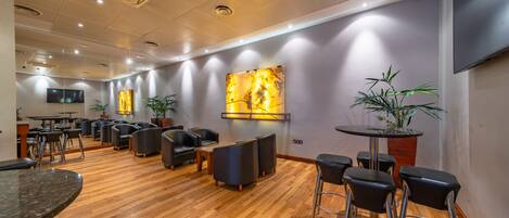 Une pièce spacieuse dotée d’un plancher en bois, de plusieurs coins salon comprenant des tables rondes et des tabourets de bar, d’un téléviseur à écran plat fixé au mur et de plantes en pot.