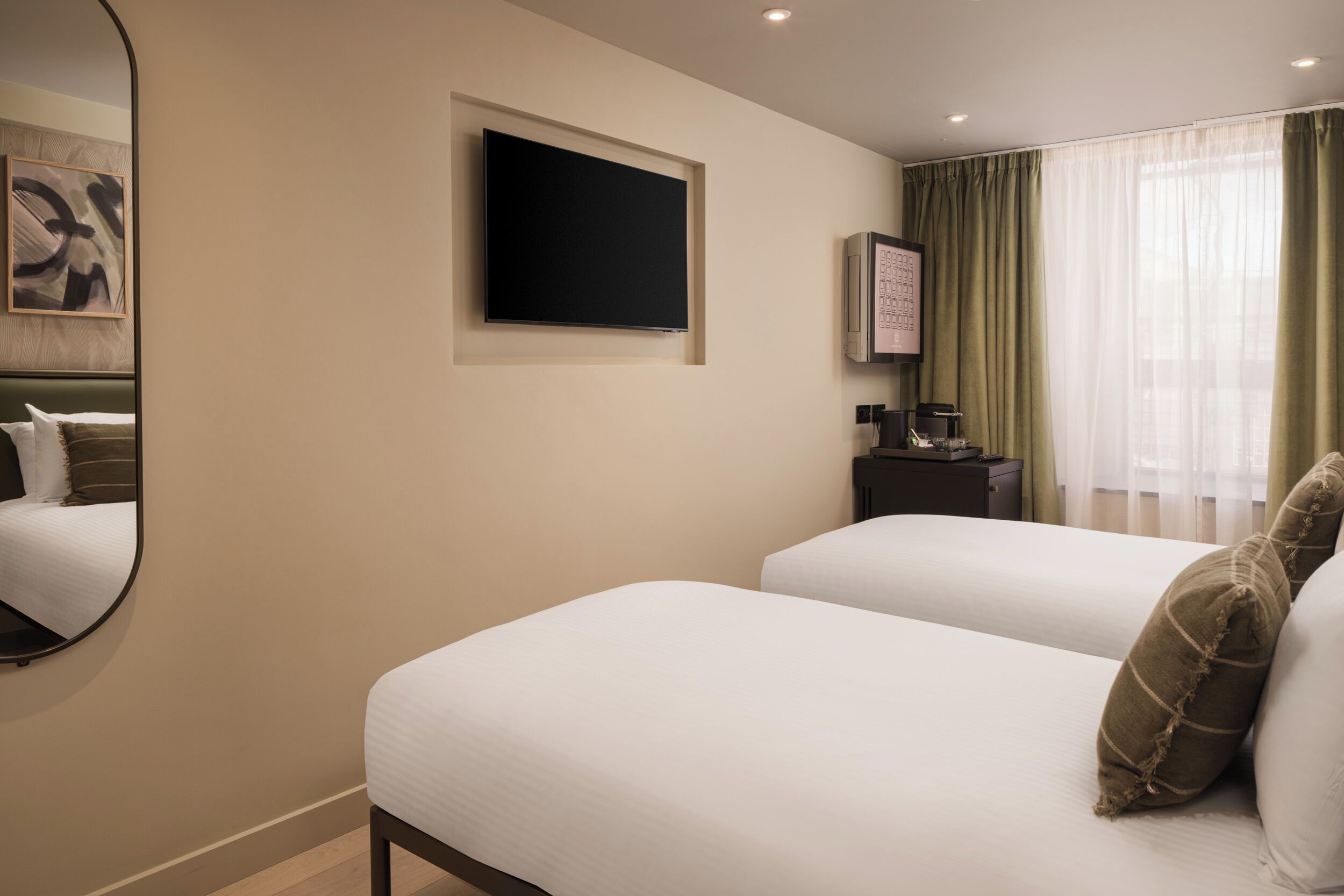 A hotel room with two beds, a flat-screen TV, a small table with a coffee maker, and a window with sheer curtains.