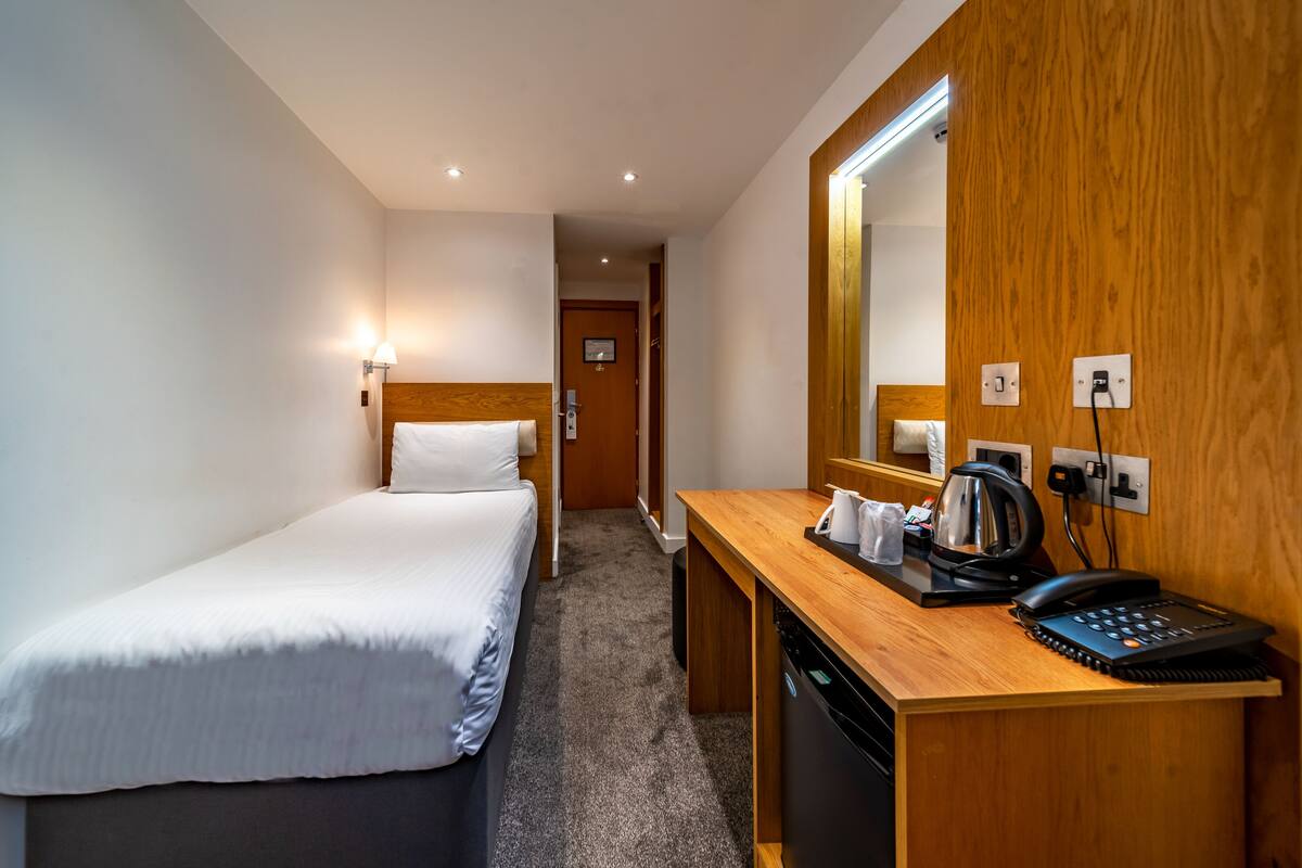 A hotel room with a single bed, a wooden desk with a telephone and a kettle, and a mirror on the wall.