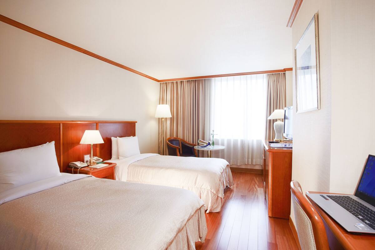 Habitació d'hotel amb dos llits, un escriptori amb un ordinador portàtil, una cadira i una taula petita amb una làmpada.