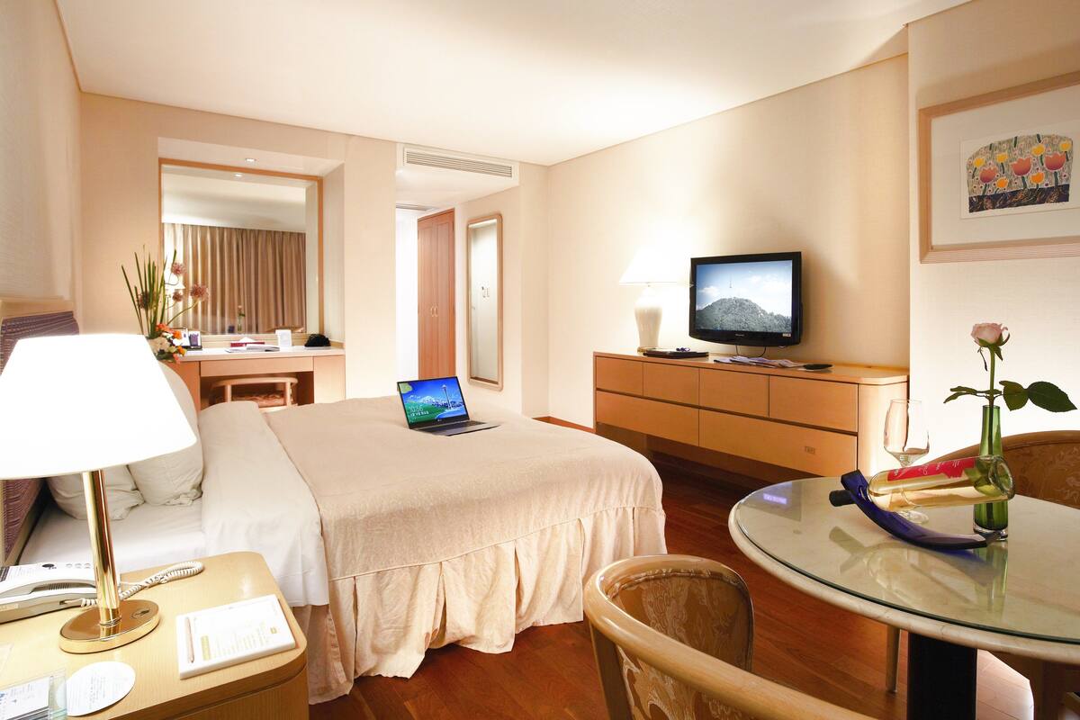Habitació d'hotel amb un llit, una taula de nit amb una làmpada, un escriptori amb un ordinador portàtil, una televisió, una taula de menjador amb un vas i una rosa, i una cadira.