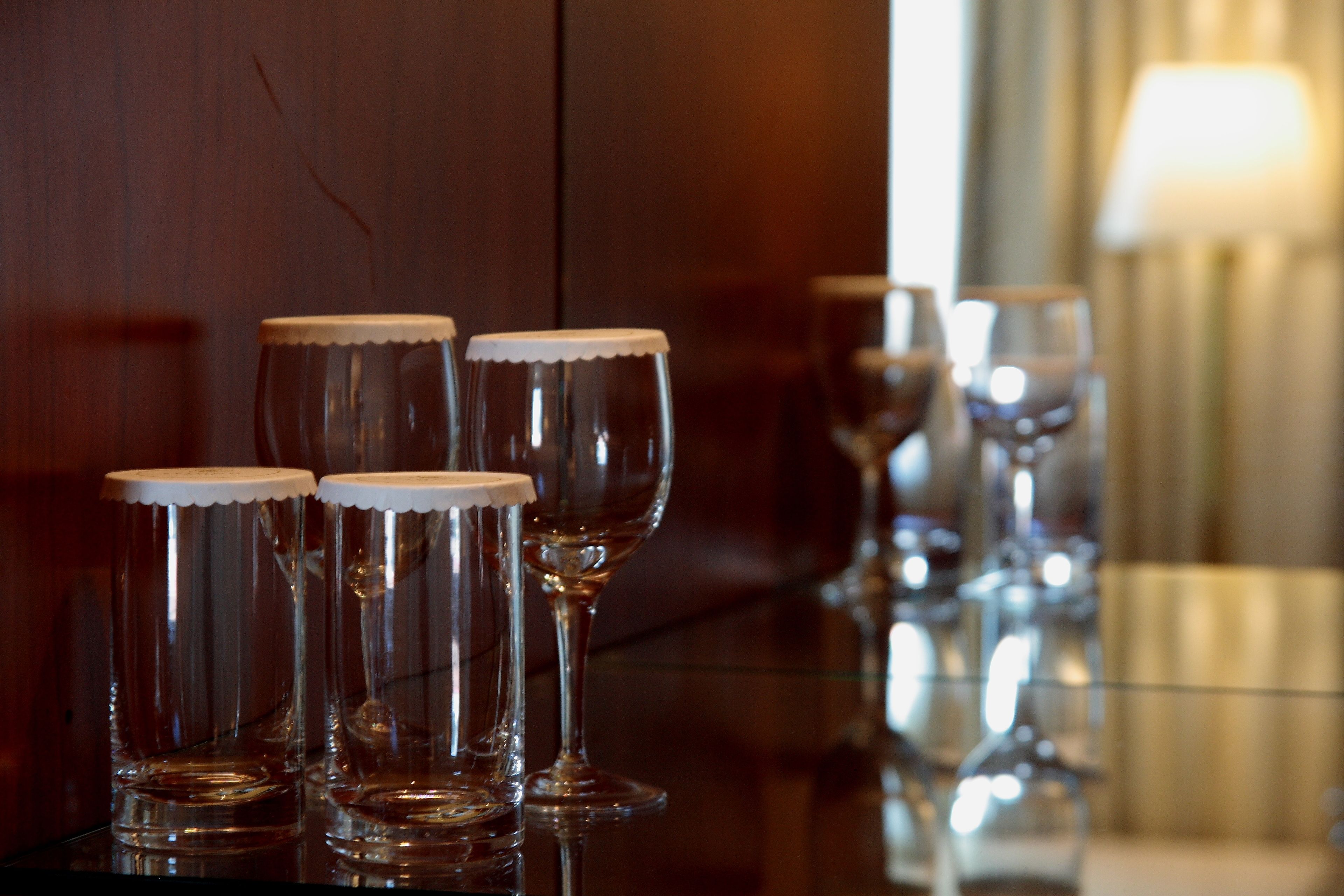 A row of empty wine glasses on a reflective surface, with a wooden cabinet and a lamp in the background.