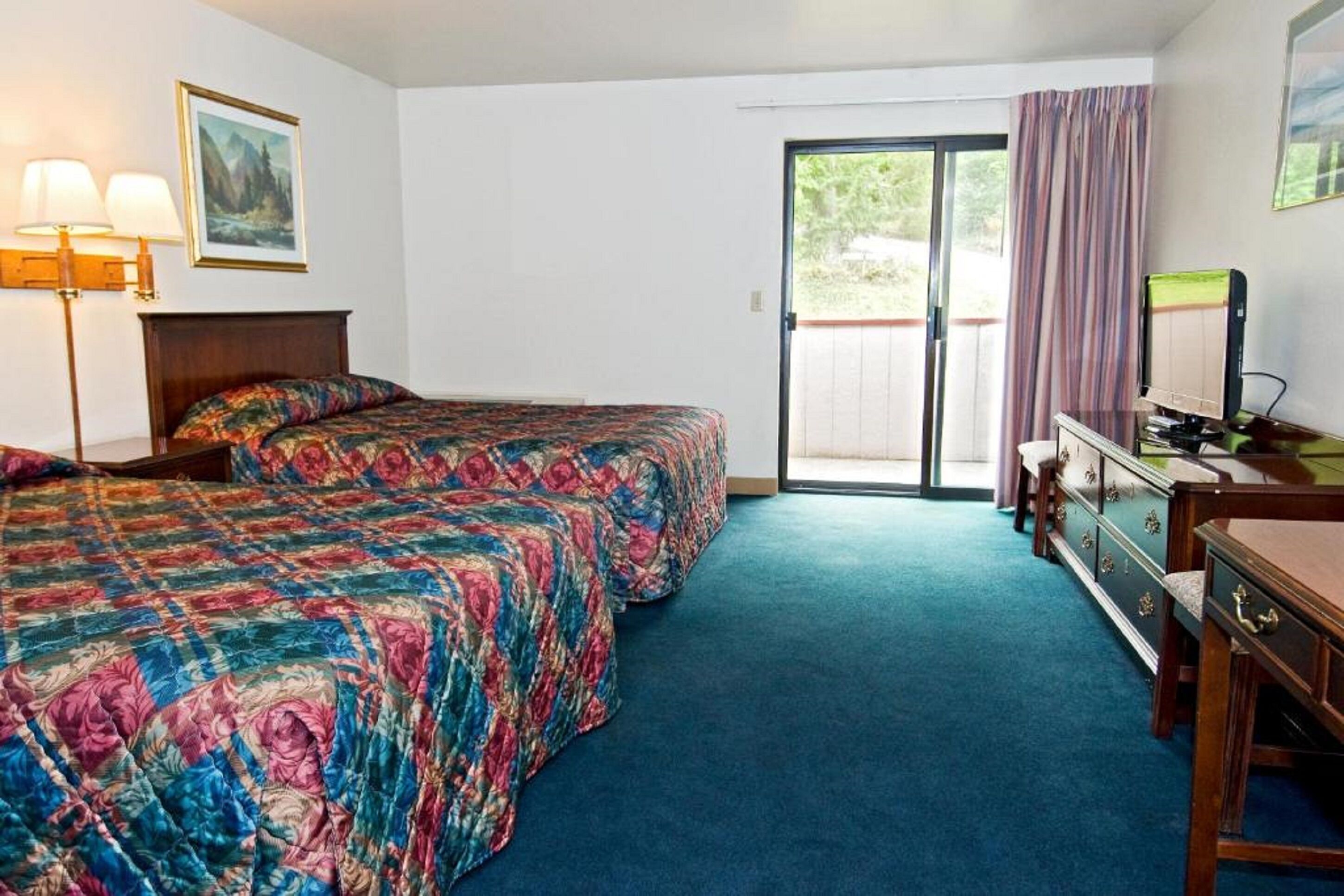 Ett hotellrum med en stor säng, ett skrivbord med en dator, en byrå och en skjutdörr i glas som leder ut till utsidan.
