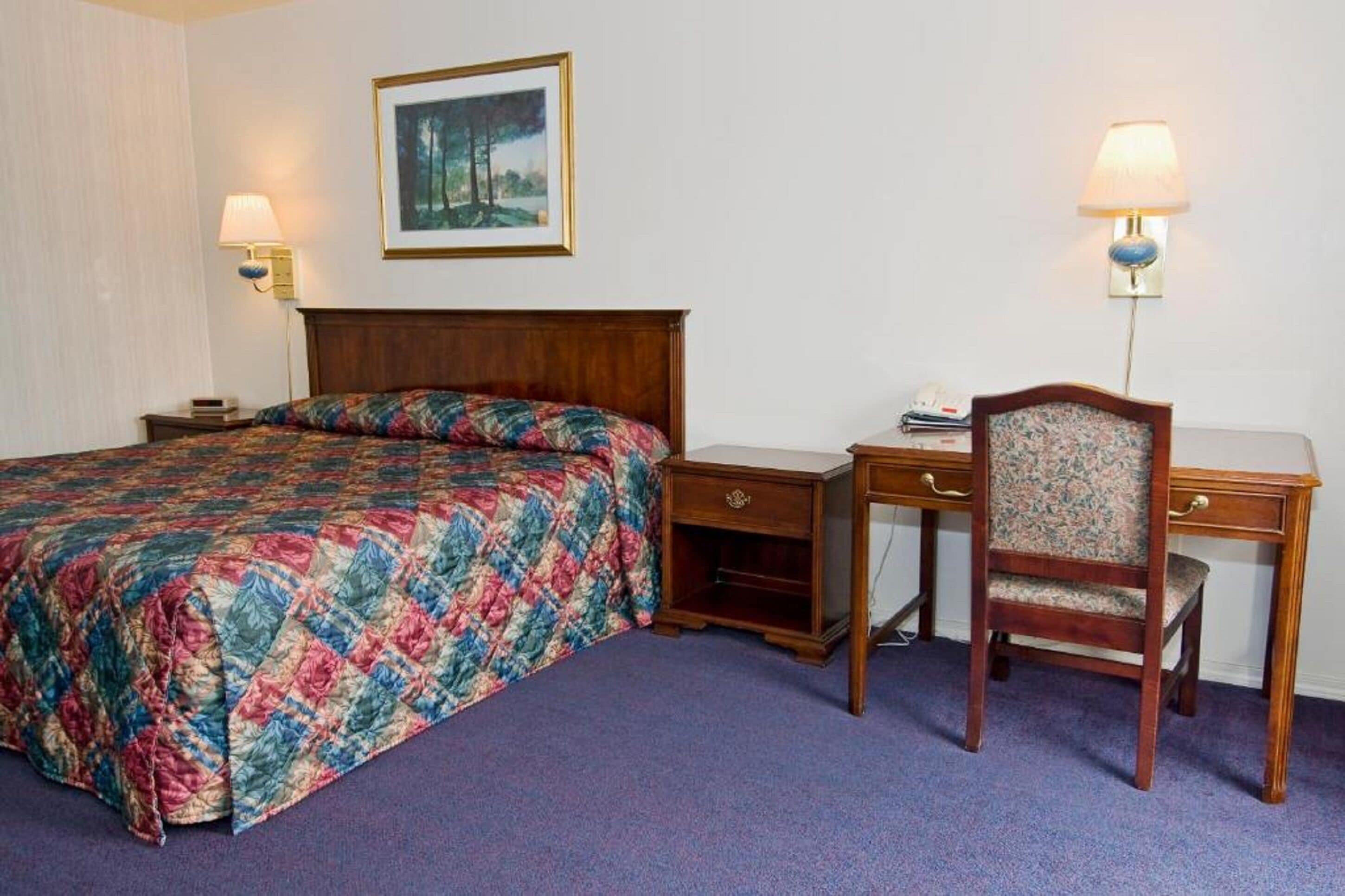 Quarto de hotel com uma cama, uma escrivaninha, dois mesinhas de cabeceira e uma cadeira.