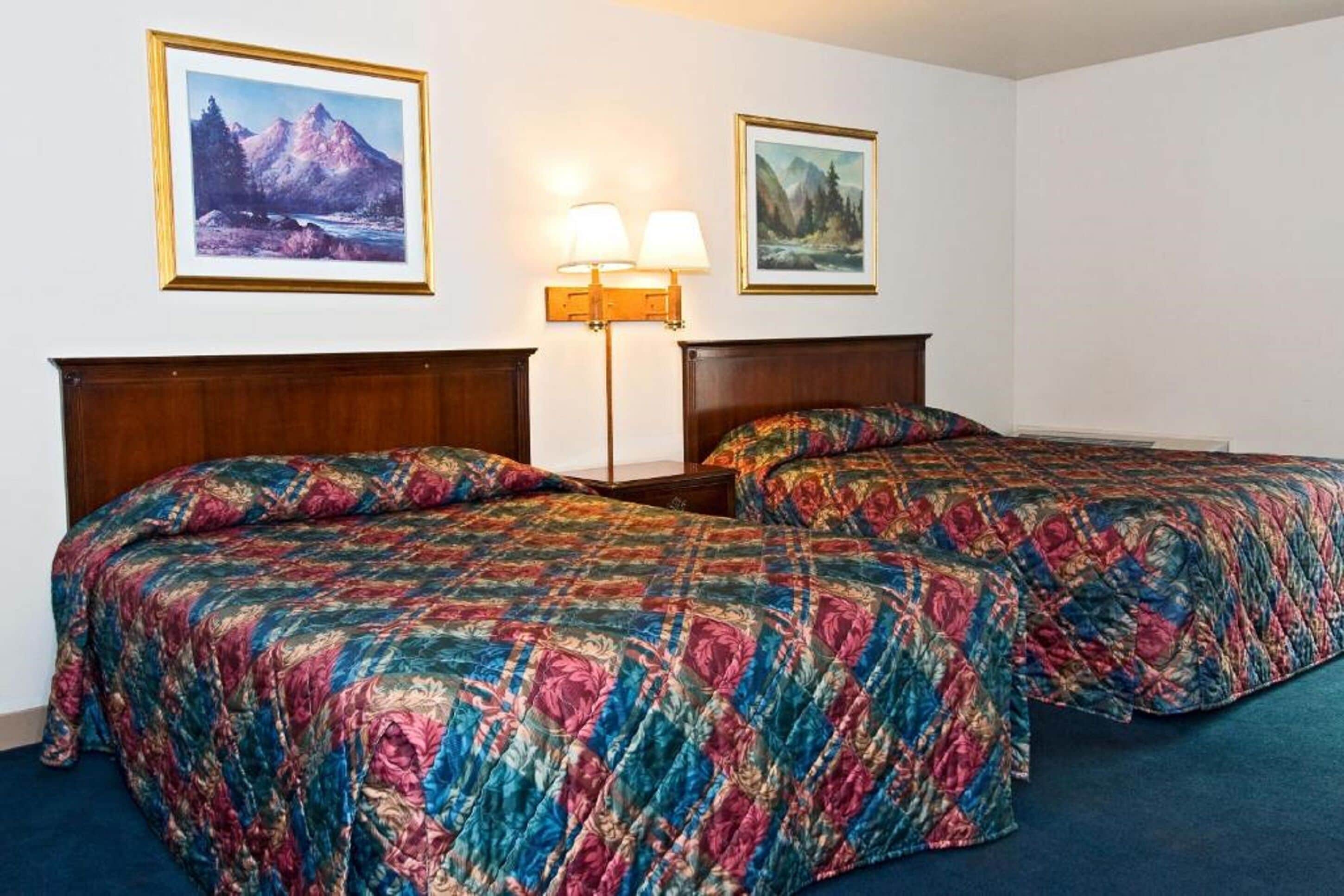 A hotel room with two beds, each with a floral-patterned bedspread, a nightstand with a lamp, and two framed landscape paintings on the wall.