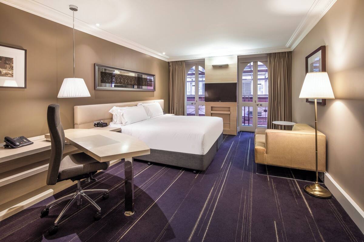 A hotel room with a bed, desk, chair, TV, and a balcony with curtains.