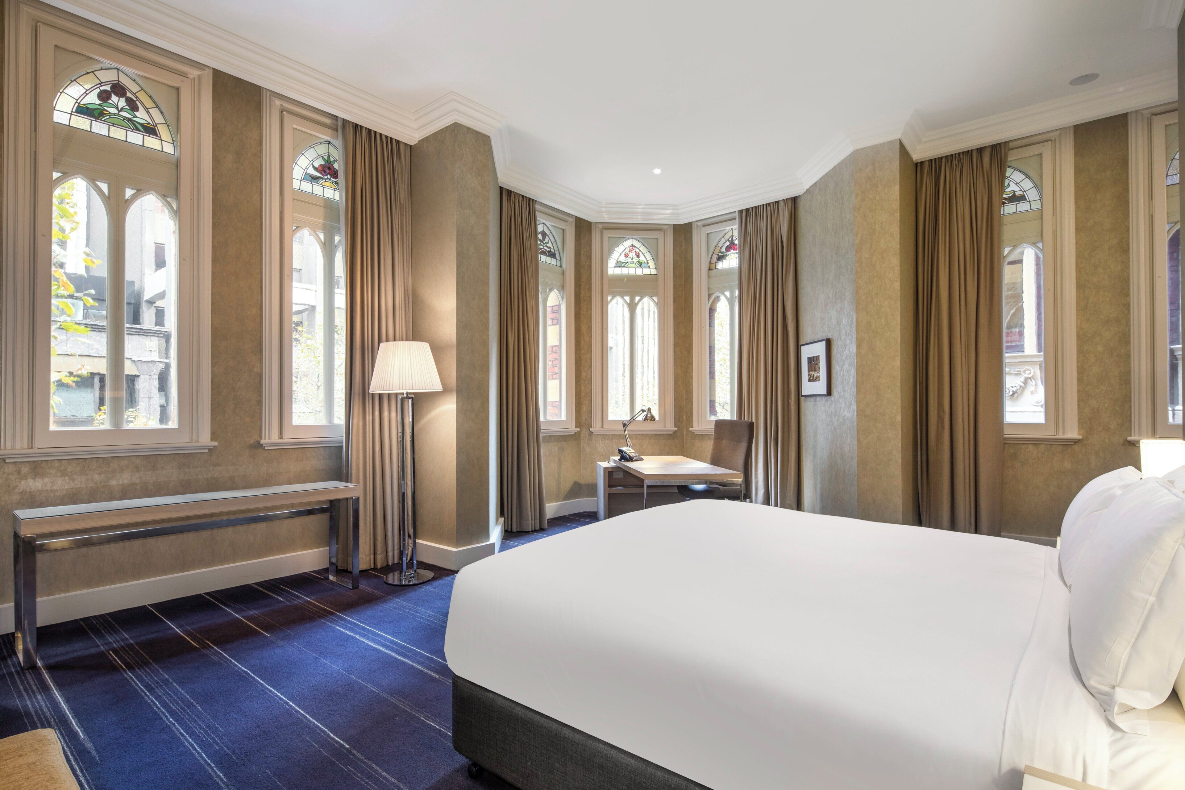 A hotel room with a large bed, a desk, a chair, and a bench. There are stained glass windows and curtains.