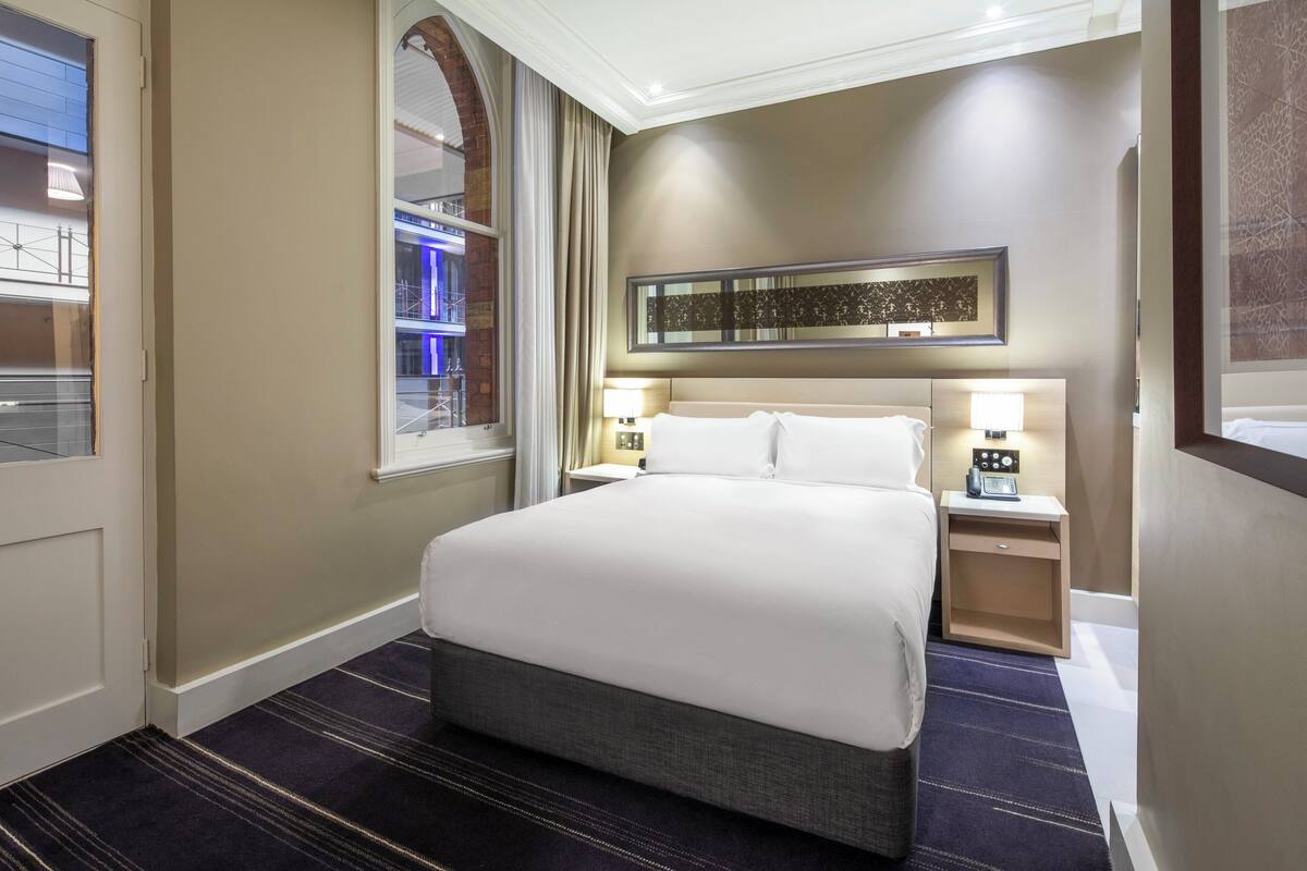 A hotel room with a large bed, two bedside tables with lamps, a window with curtains, and a wall-mounted artwork.