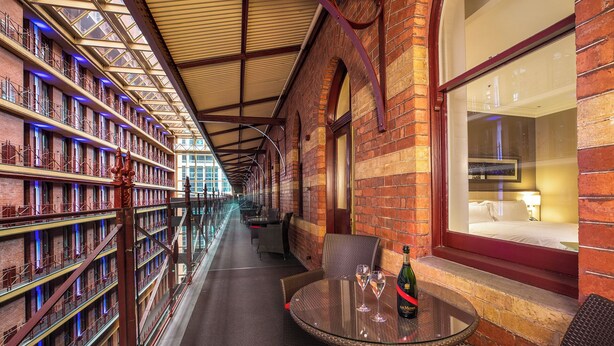 A corridor with a glass railing, a table with two glasses and a bottle, and a bed visible through a window. - InterContinental Melbourne The Rialto by IHG (Melbourne)