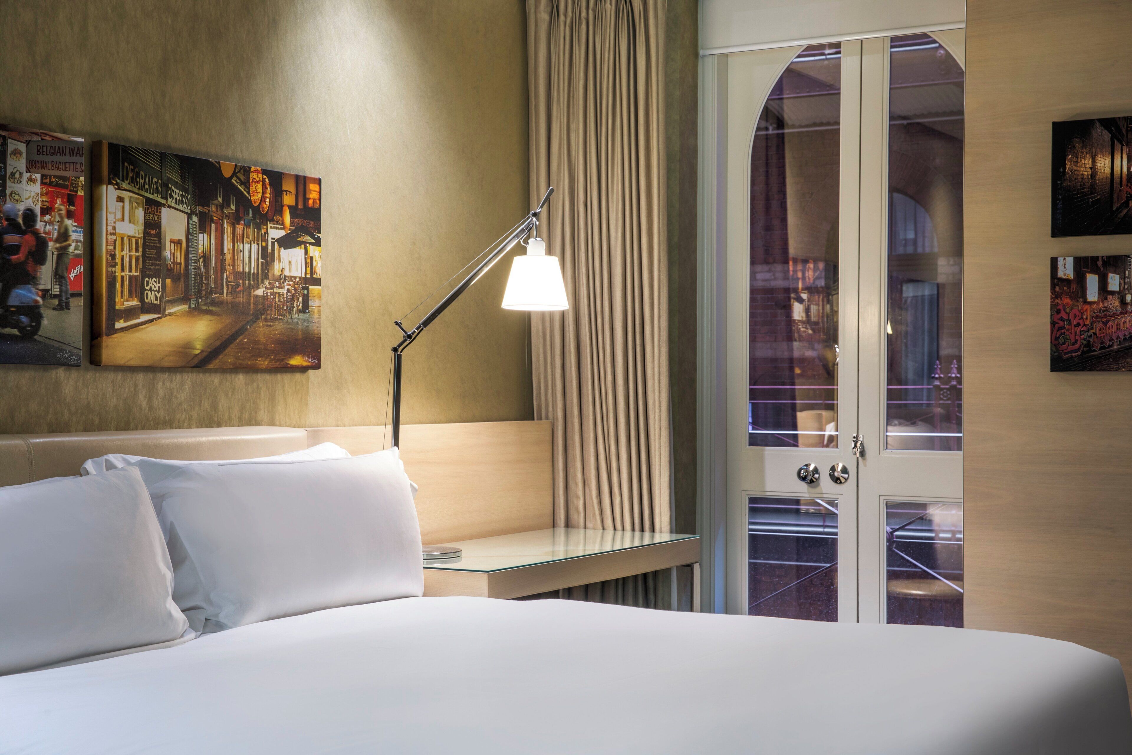 A hotel room with a bed, a bedside table, a lamp, a window with curtains, and artwork on the wall.
