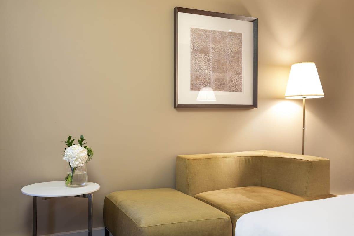 Habitación de hotel con una cama, una pequeña mesa redonda con un florero y una floristería, un cuadro en la pared y una lámpara de pie.
