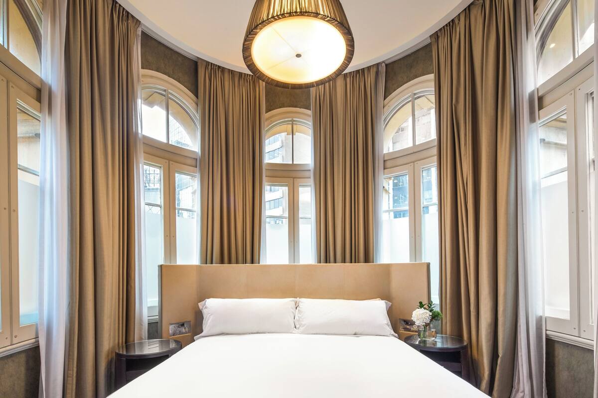 A large bed with a wooden headboard, flanked by curtains, in a room with large windows and a ceiling light fixture.