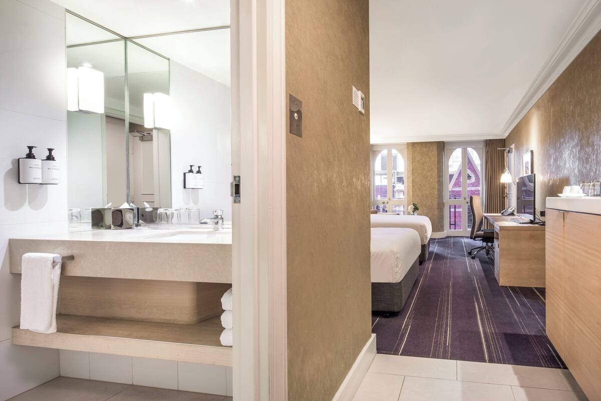 A hotel room with a large mirror, a vanity with toiletries, a bed, a desk, and a view of a building with arches.