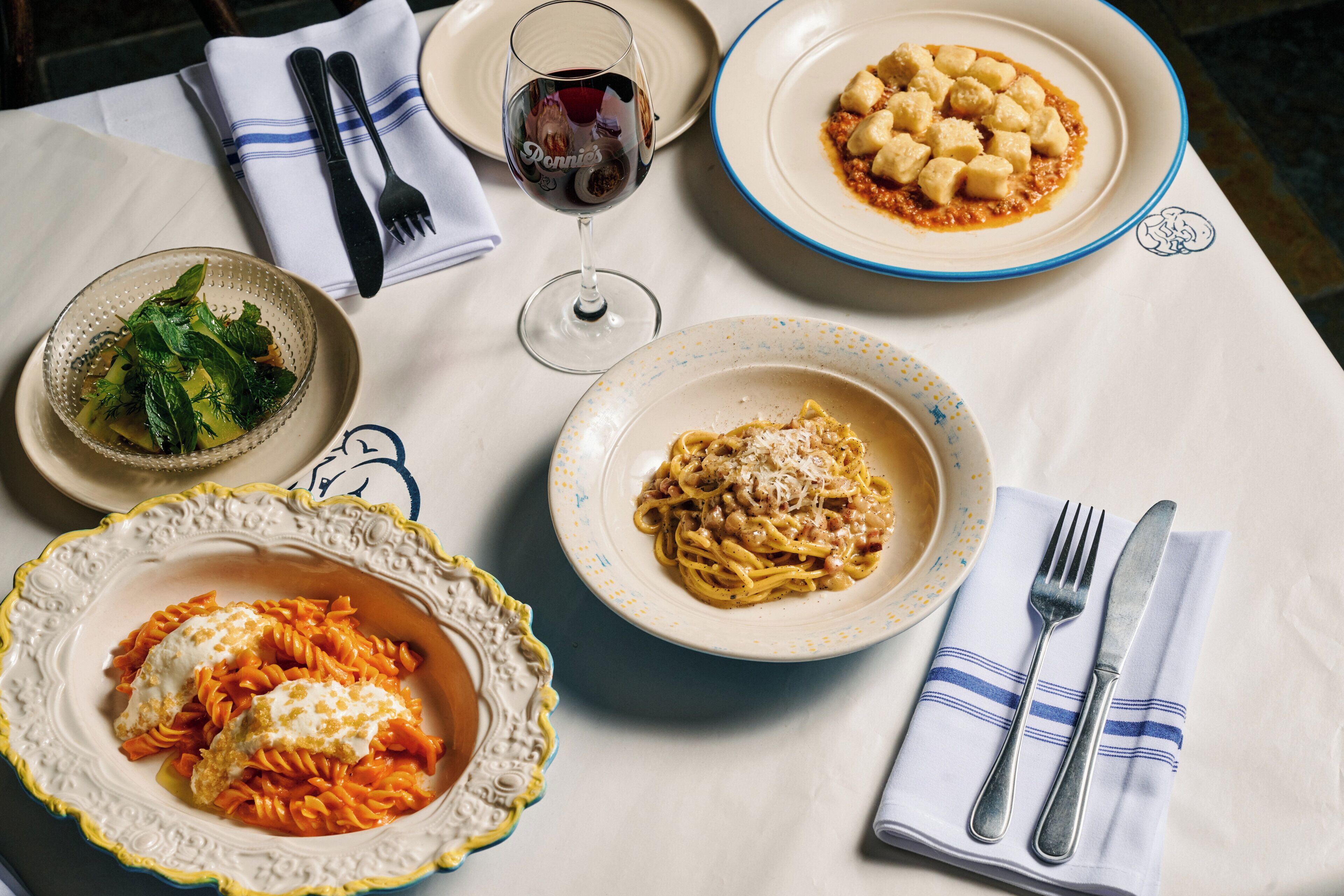 A table with various dishes and cutlery, including pasta and a glass of red wine.